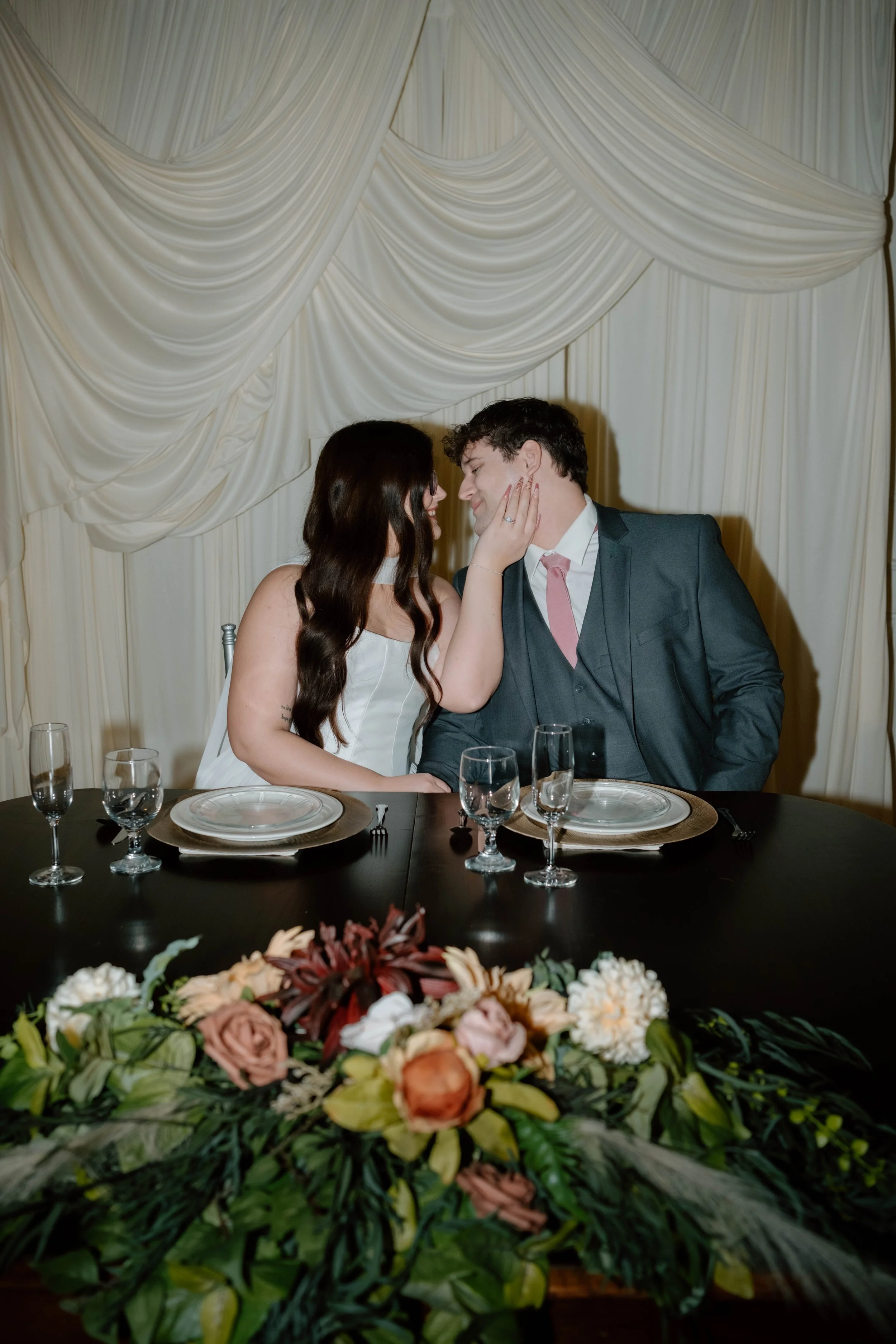 A couple sitting at a decorated dinner table, touching faces affectionately, with a floral centerpiece in the foreground and draped curtains in the background.