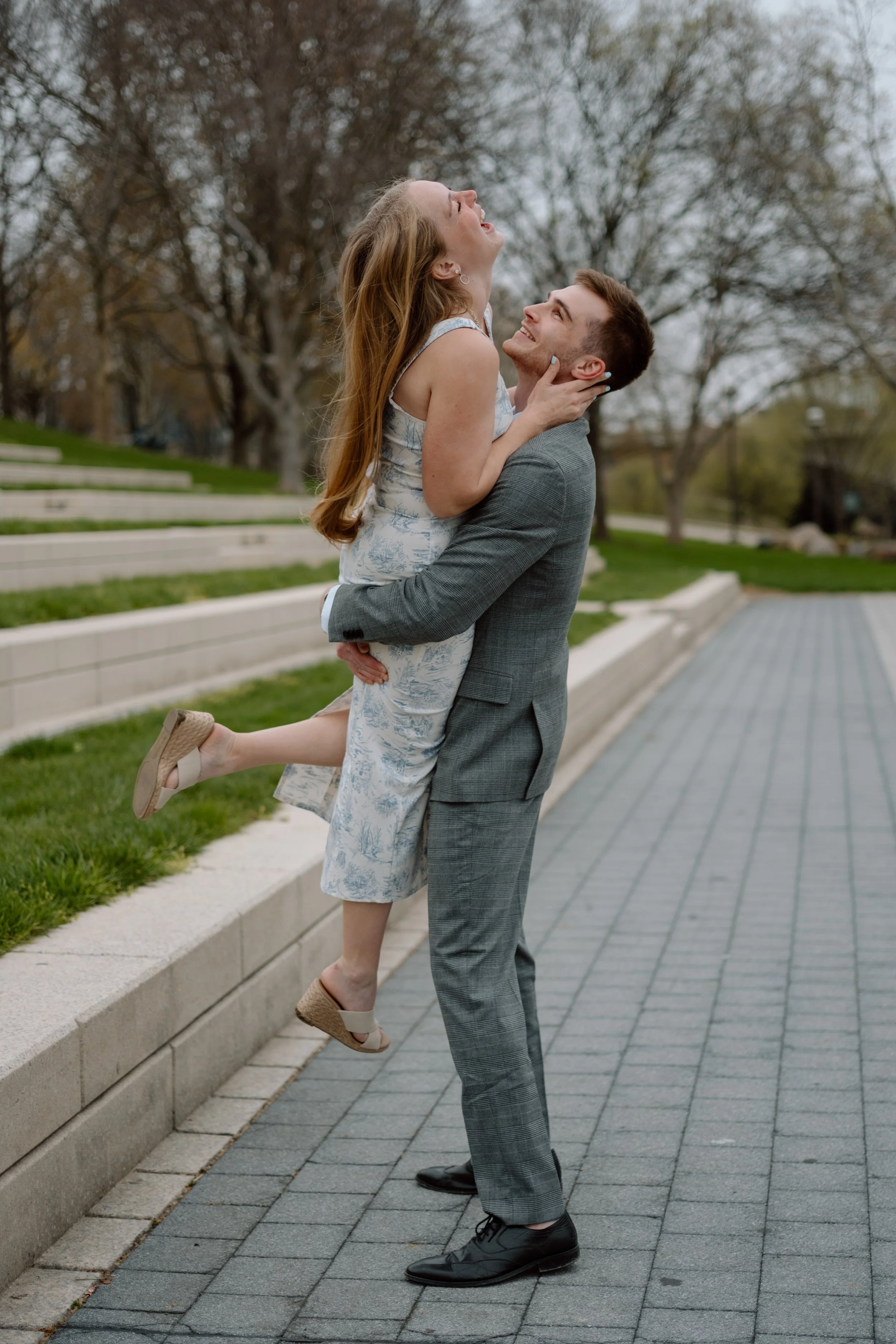 A man in a gray suit lifts a woman in a sleeveless, floral dress in an outdoor park, both smiling and looking happy. She has long, red hair and is wearing beige wedge sandals. The scene is set in an area with steps and trees.