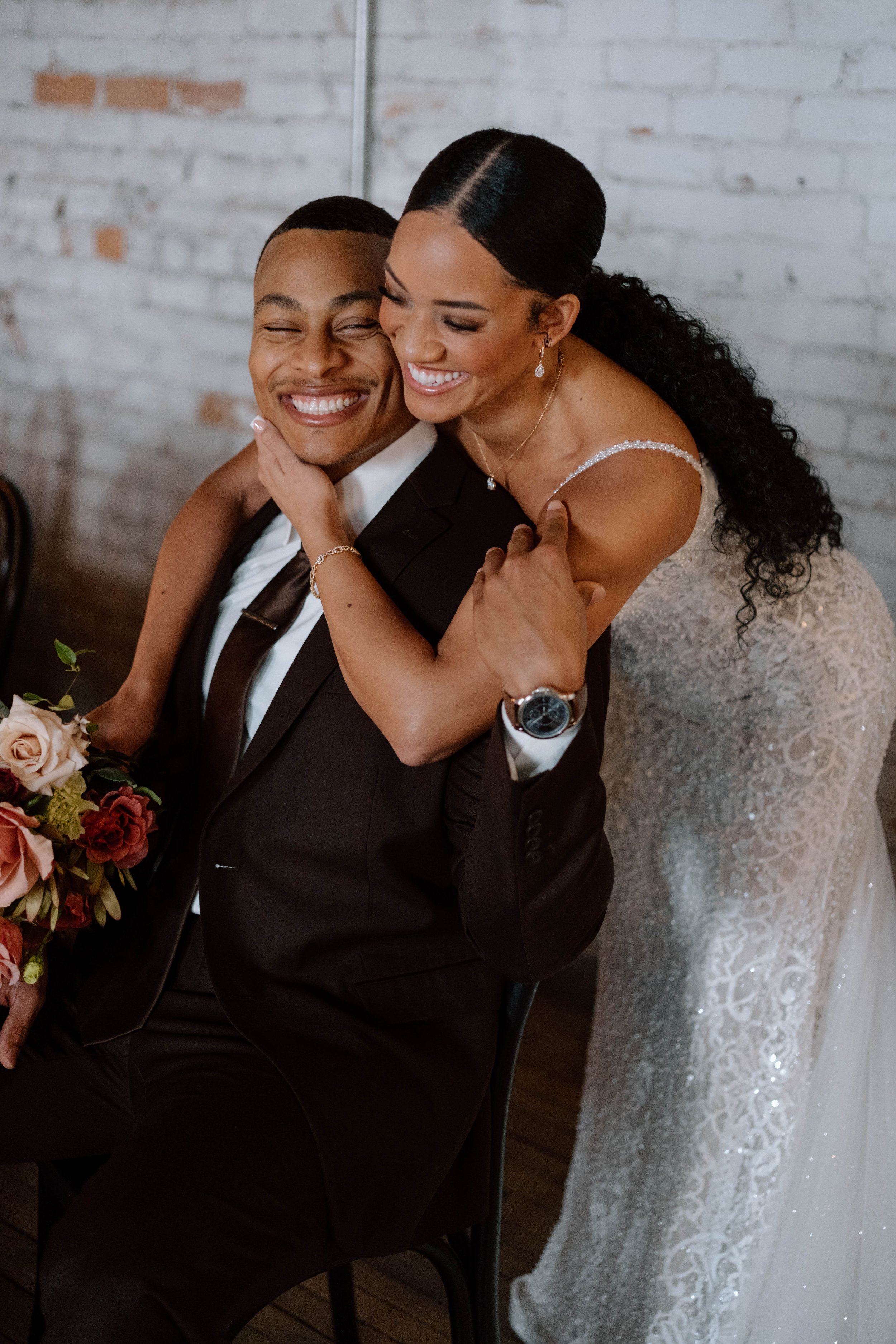 A joyful couple dressed in wedding attire, with the woman hugging the seated man who is smiling, holding a bouquet of flowers, against a white brick wall background.