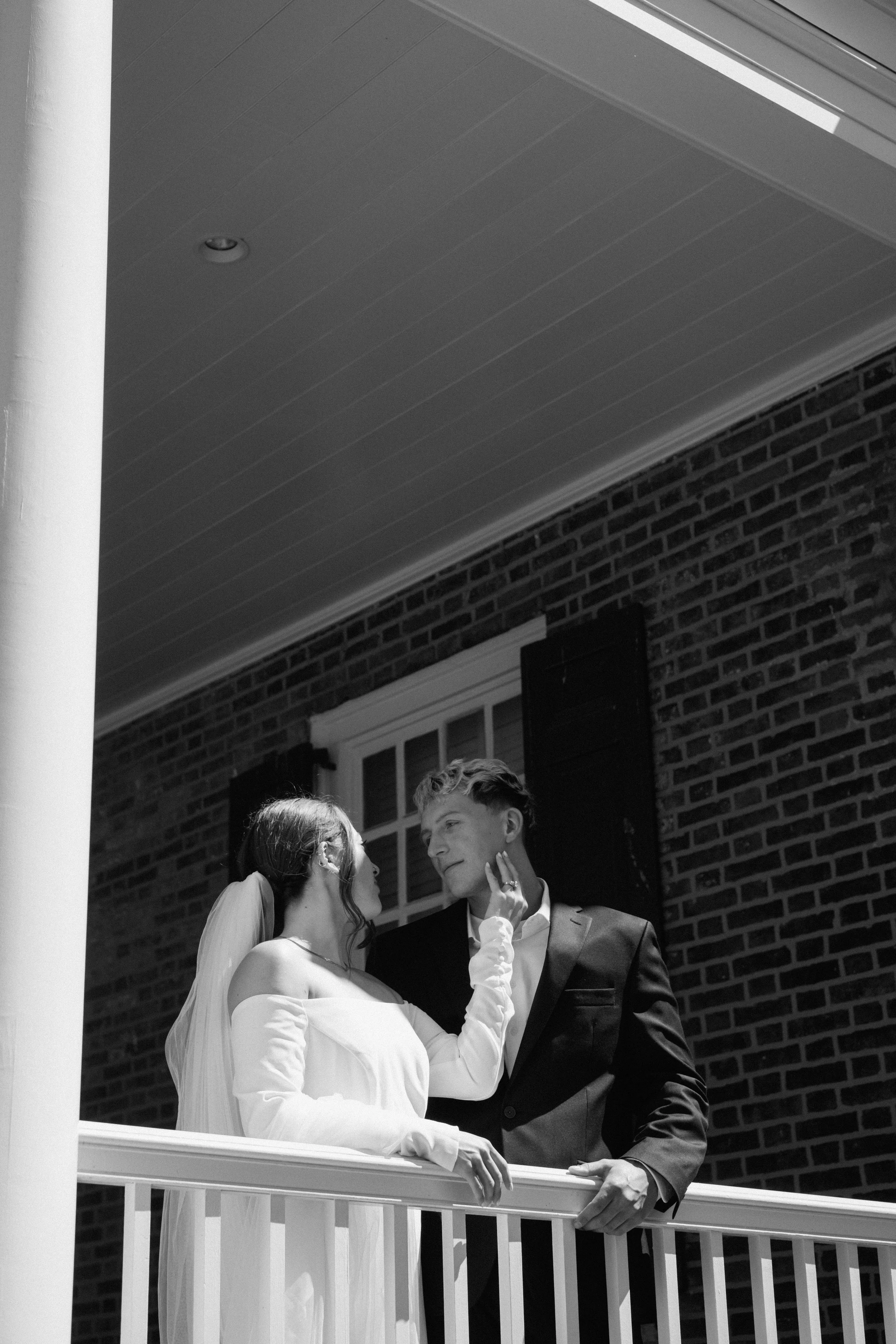 Black and white photo of a bride and groom on a porch, gazing at each other. The bride with long hair and a veil touches the groom's face, who is wearing a suit. They stand in front of a brick wall and a window.