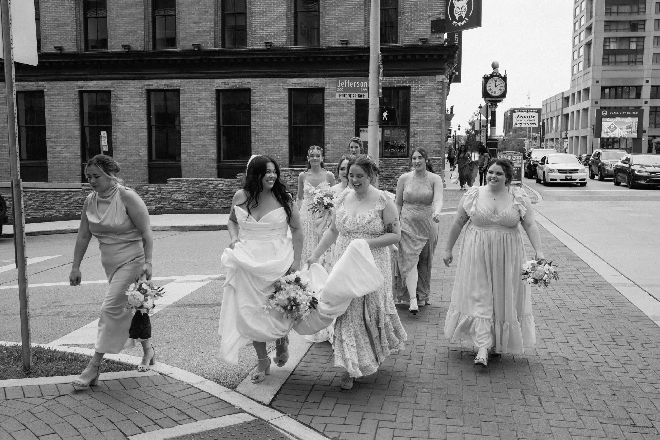 A group of women, dressed in elegant dresses, walking on a city sidewalk, holding bouquets of flowers, possibly for a wedding or special event.