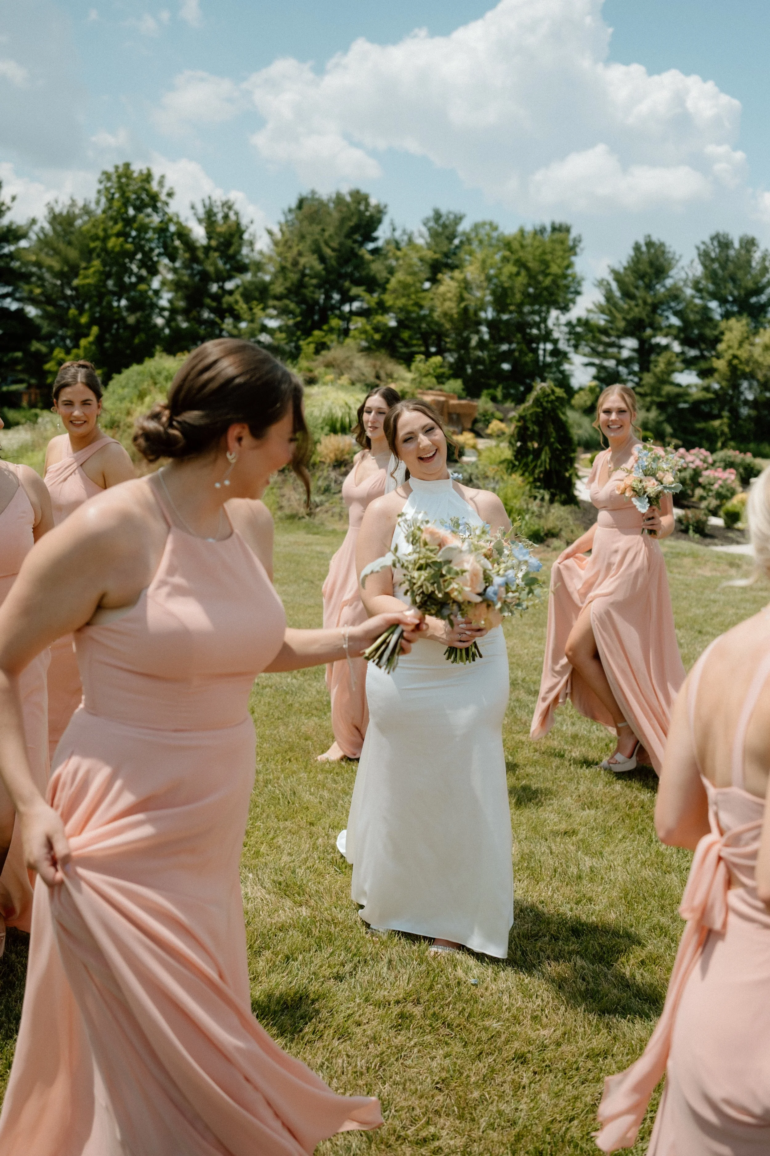 Bridal party standing outdoors on a grassy lawn, with five women dressed in pastel pink bridesmaid dresses and one woman in a white wedding gown, holding bouquets of flowers, smiling and enjoying the sunny day with trees and a partly cloudy sky in th