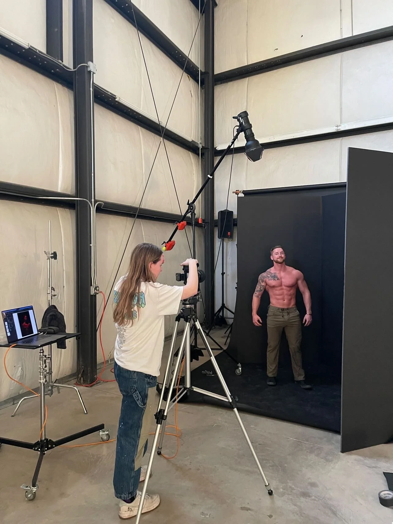 A woman is taking a professional photo of a muscular man with tattoos, posing shirtless in a photography studio with a black backdrop.