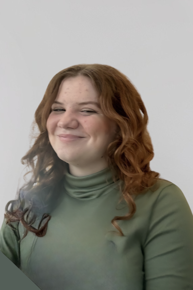 A woman with curly red hair wearing a green turtleneck, smiling with a hint of mischief in her eyes, standing against a plain white background.