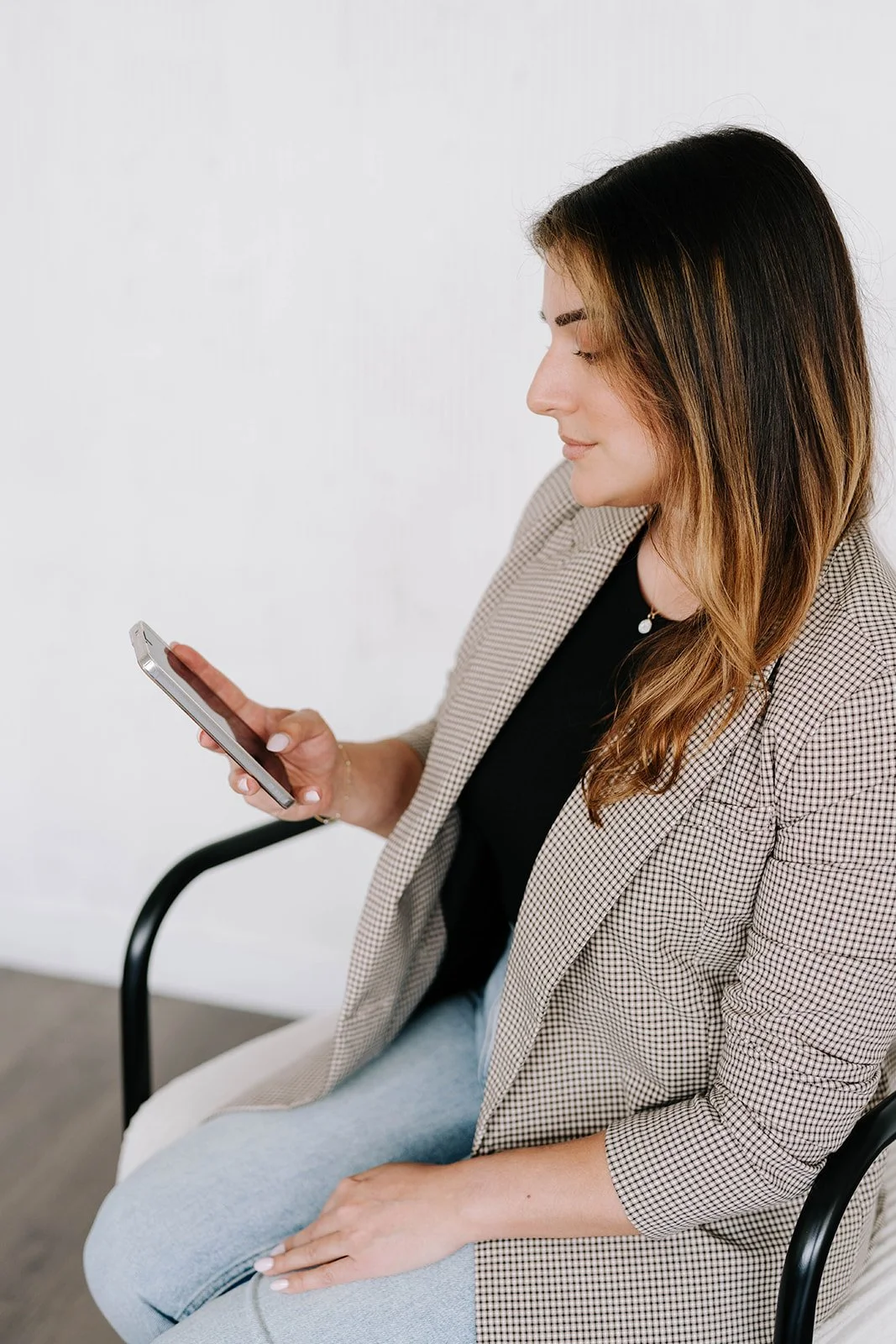 A woman sitting in a chair, looking at her smartphone, wearing a beige checkered blazer and black top, with long, wavy hair.