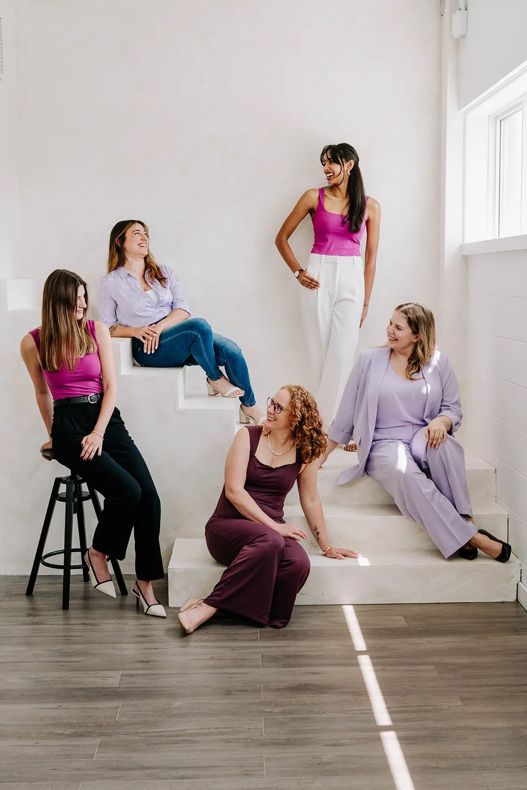 Six women in colorful outfits smiling and chatting in a bright, white room with natural light.