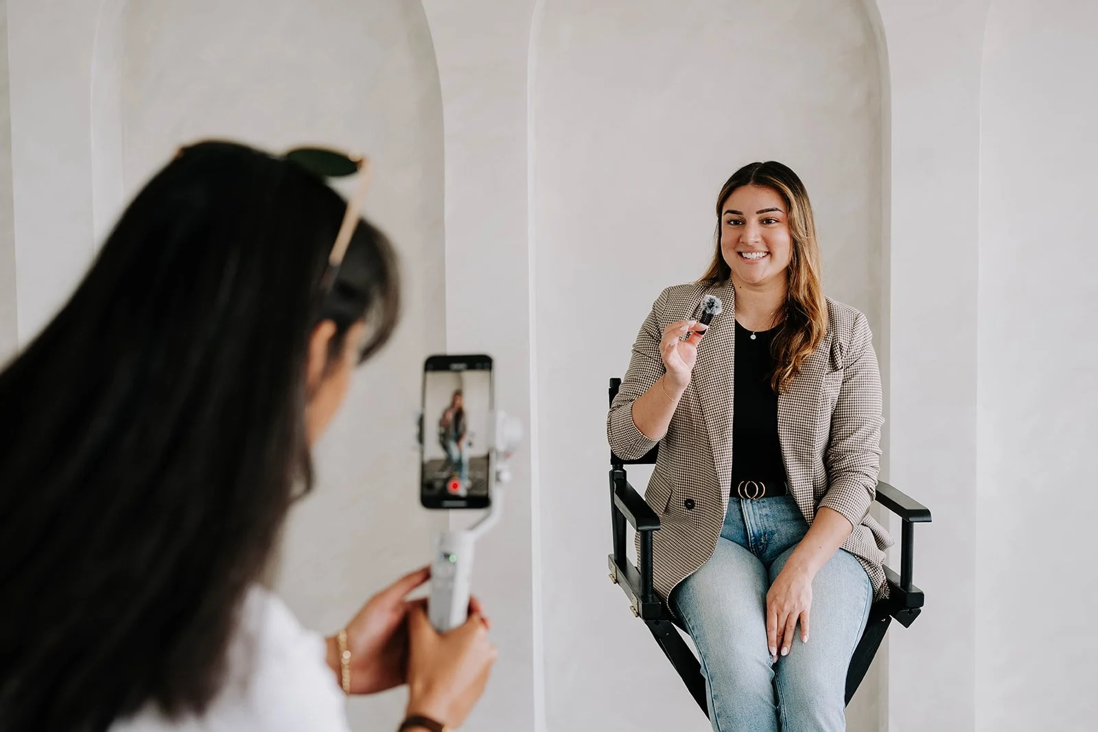 A woman in a blazer sitting on a director's chair being filmed by another woman recording with a smartphone on a stabilizer. The filming is indoors against a white wall with arches.