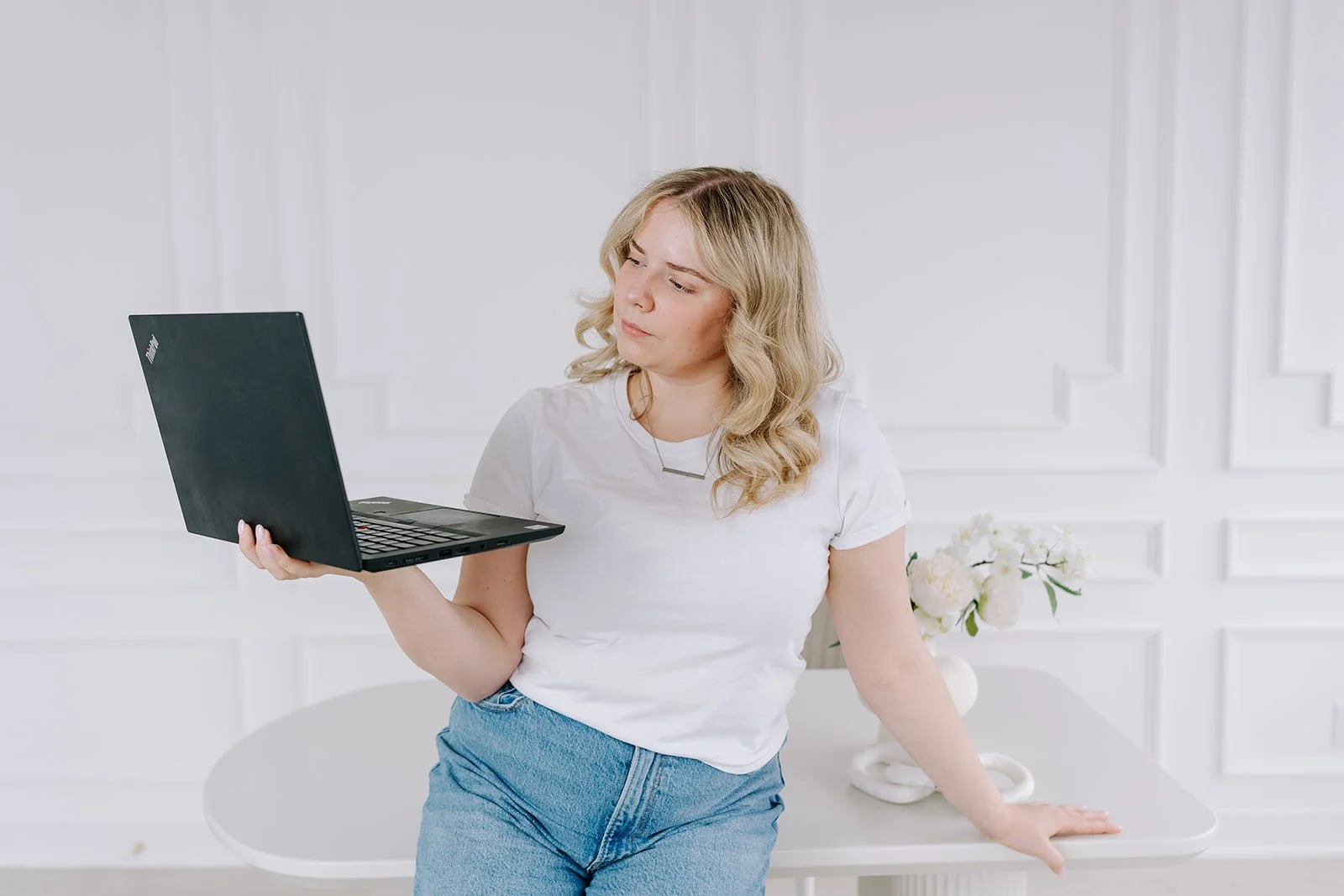 A young woman with blonde hair in loose waves, wearing a white T-shirt and blue jeans, stands near a white table with a vase of white flowers. She is holding a black laptop in her right hand and looking at it thoughtfully, in a bright, minimalist room with white paneled walls.
