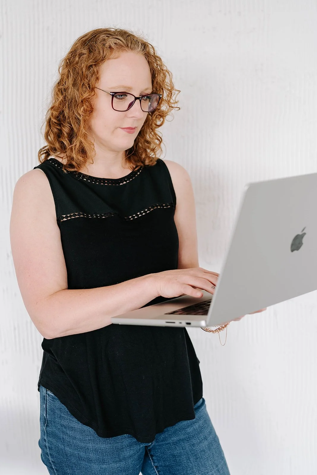 A woman with curly red hair and glasses looking at a silver MacBook laptop against a plain white wall.