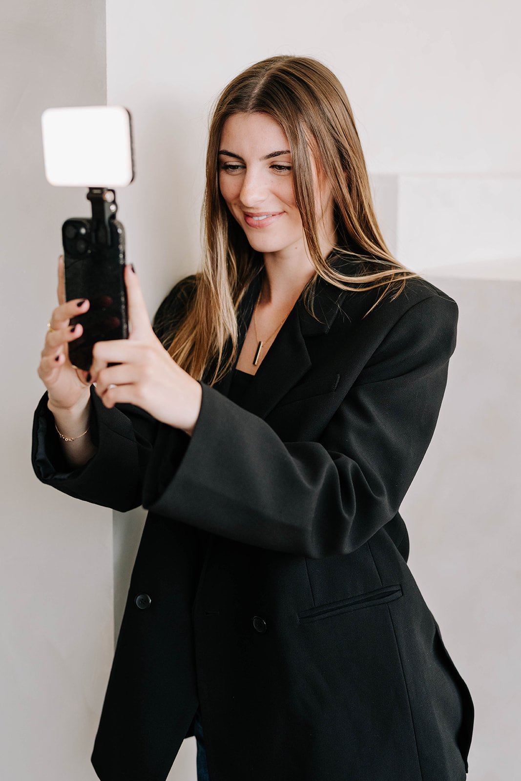 A woman with long on brown hair wearing a black blazer is taking a selfie with her smartphone.