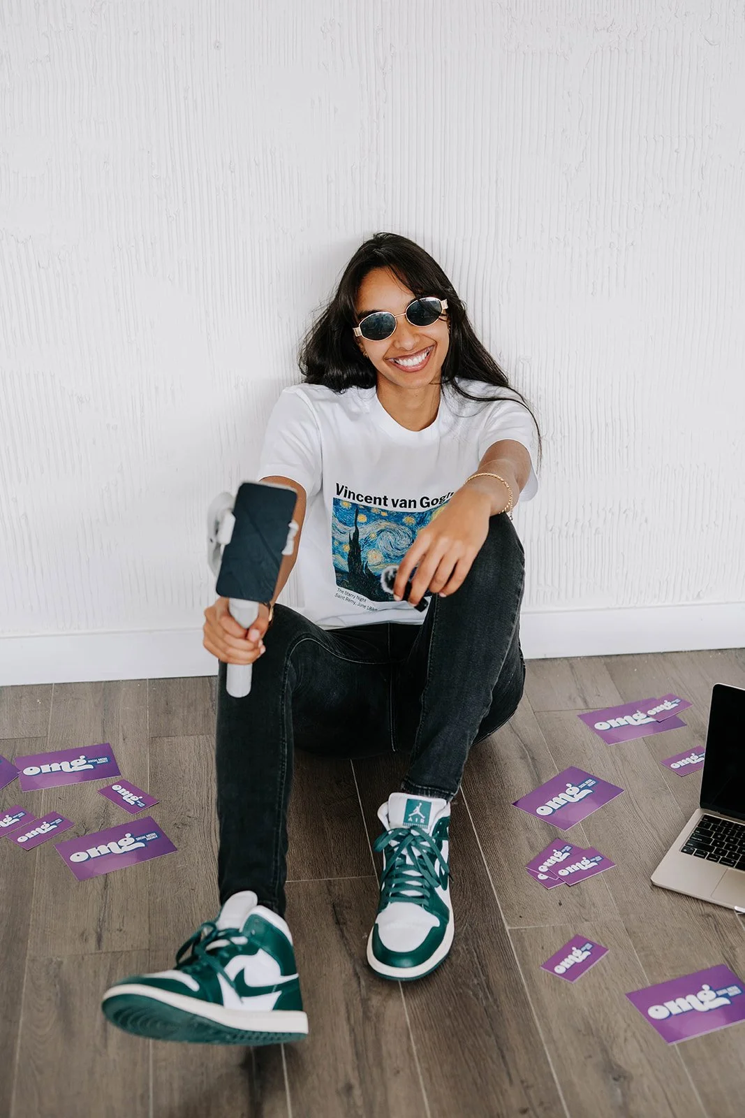 Young woman sitting on the floor, wearing sunglasses, a white t-shirt, black jeans, and teal and white sneakers, holding a selfie stick. Around her on the floor are several purple cards with the word 'Omie' on them, and a laptop is partially visible on the right side.