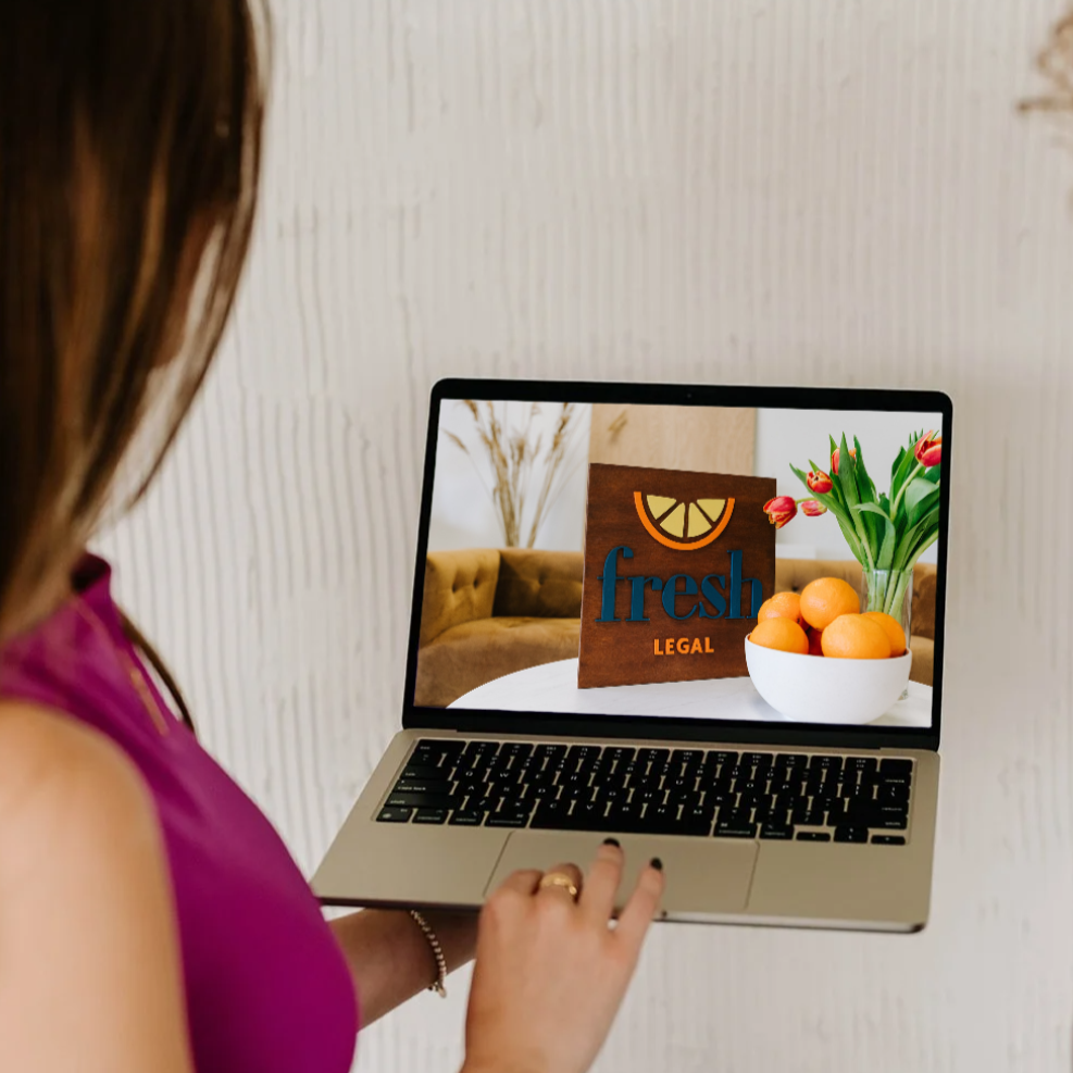 Person holding a laptop with a screen displaying a wooden sign with a lemon wedge, the word 'fresh', and 'LEGAL', along with a bowl of oranges and a vase of pink tulips in the background.