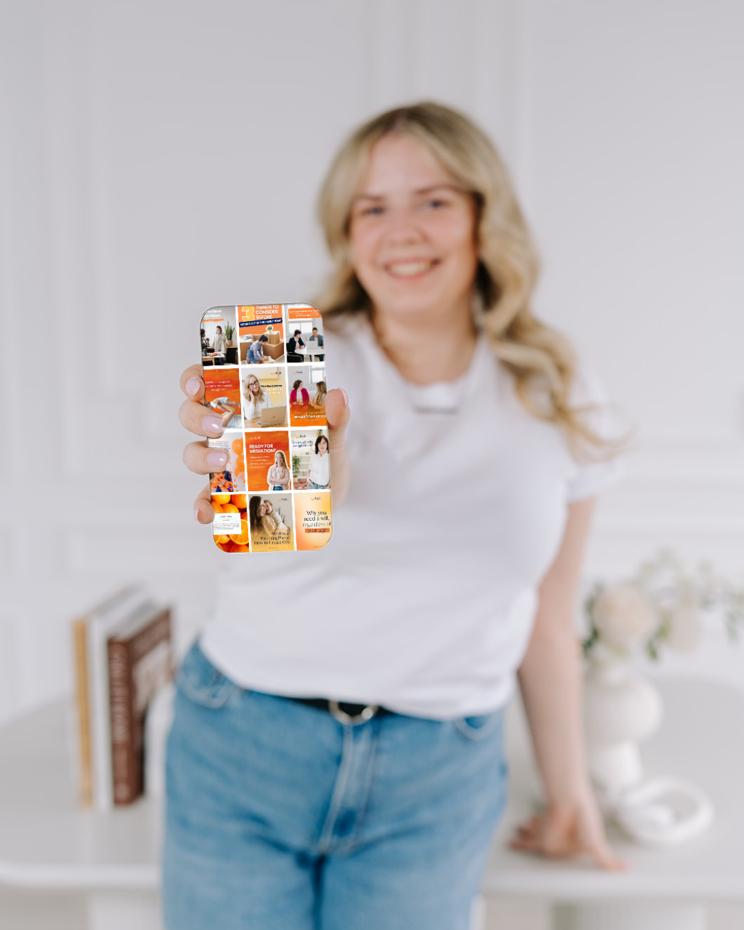 A woman with blonde hair in a white t-shirt and blue jeans is smiling and holding a smartphone towards the camera, showing a colorful grid of images and text on the screen.