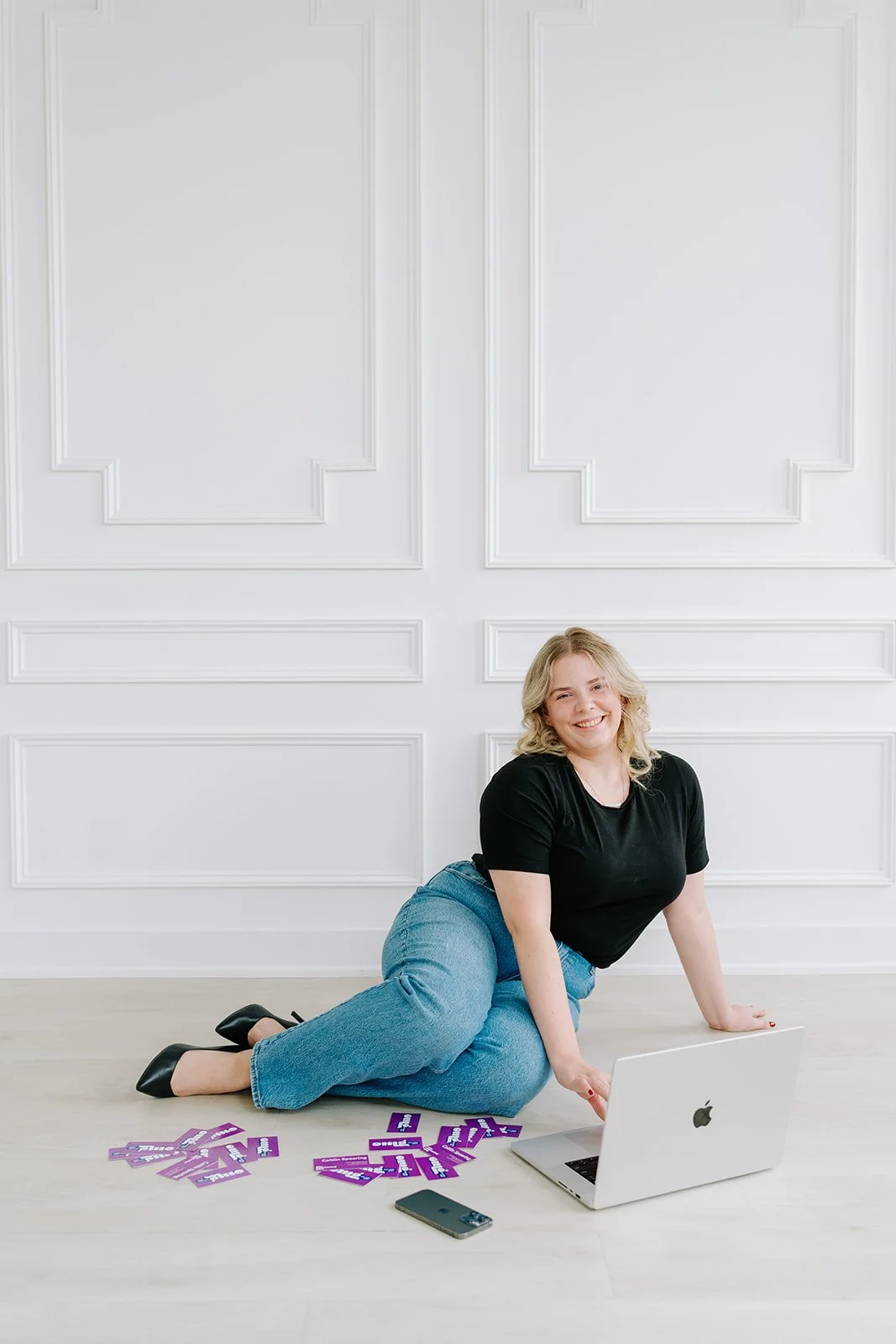 A woman with blonde hair wearing a black shirt and blue jeans, sitting on the floor with her knees bent, smiling at the camera. She is surrounded by purple cards with white and pink text, a silver laptop, and a smartphone against a white wall with decorative paneling.