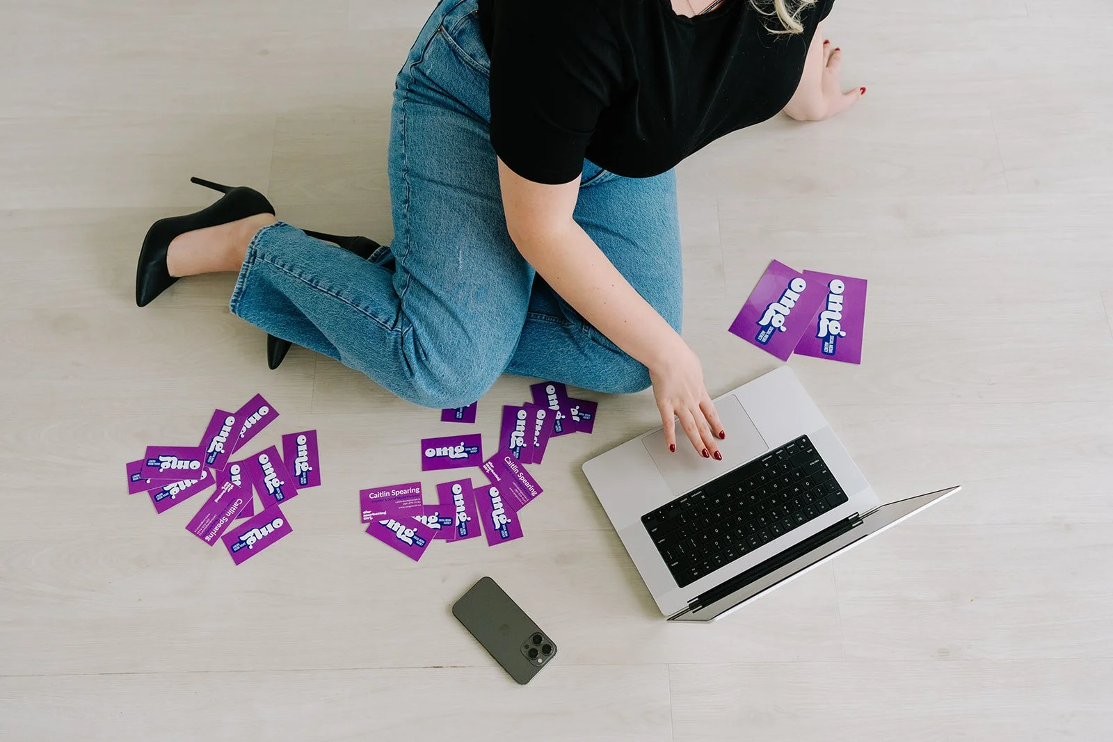 A woman is sitting on the floor with a laptop in front of her, surrounded by purple campaign flyers that read 'Olio'. She is wearing black high heels, blue jeans, and a black top, and has blonde hair. Her left hand is pointing at the laptop's touchpad, and there is a smartphone nearby.