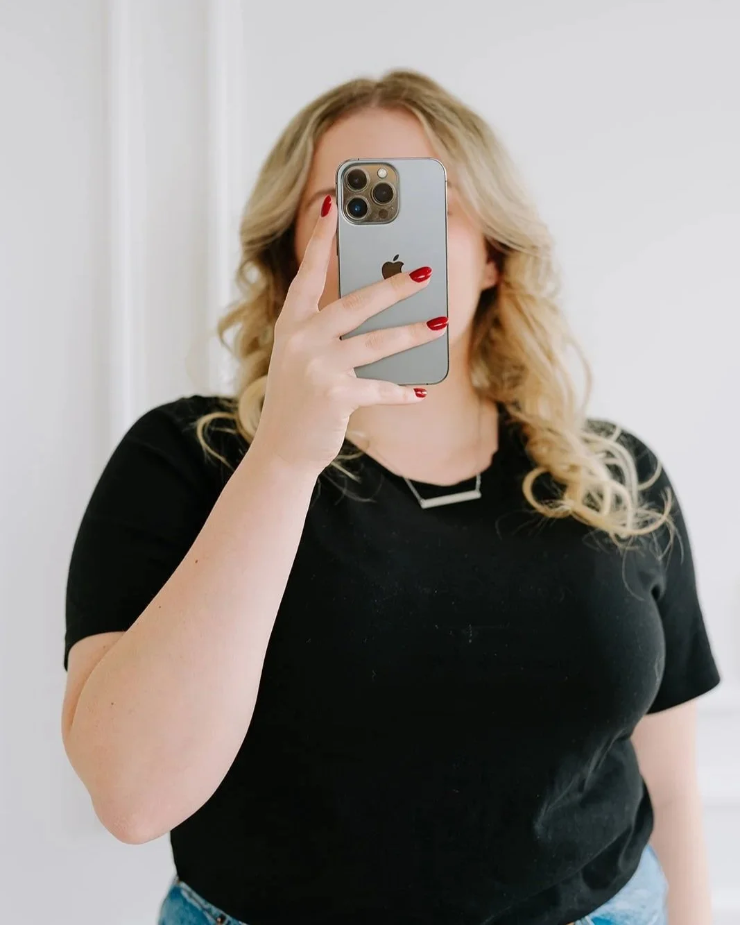 A woman with blonde curly hair taking a selfie with a smartphone, wearing a black t-shirt and red nail polish.