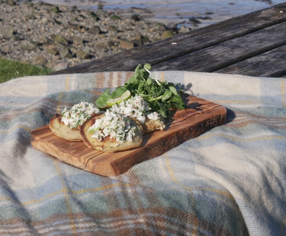 Seafood tostadas with crab meat topping, lime slices, and fresh greens on a wooden serving board outdoors near water.