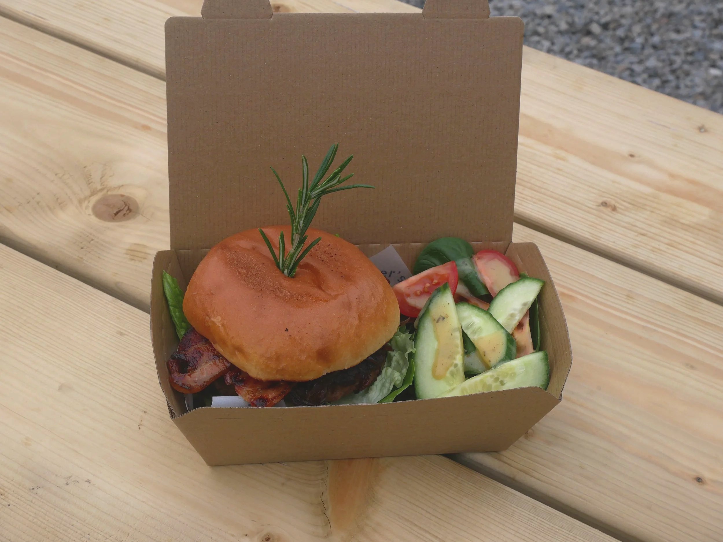 Burger with bacon and rosemary in a cardboard box, alongside cucumber and tomato salad on a wooden table.