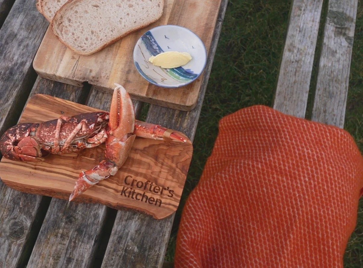 A wooden cutting board with a lobster on it, the board has 'Crofter's Kitchen' engraved on it. Next to the lobster, there is a small plate with a pat of butter. There are two slices of bread on a second wooden board. An orange textured towel is parti