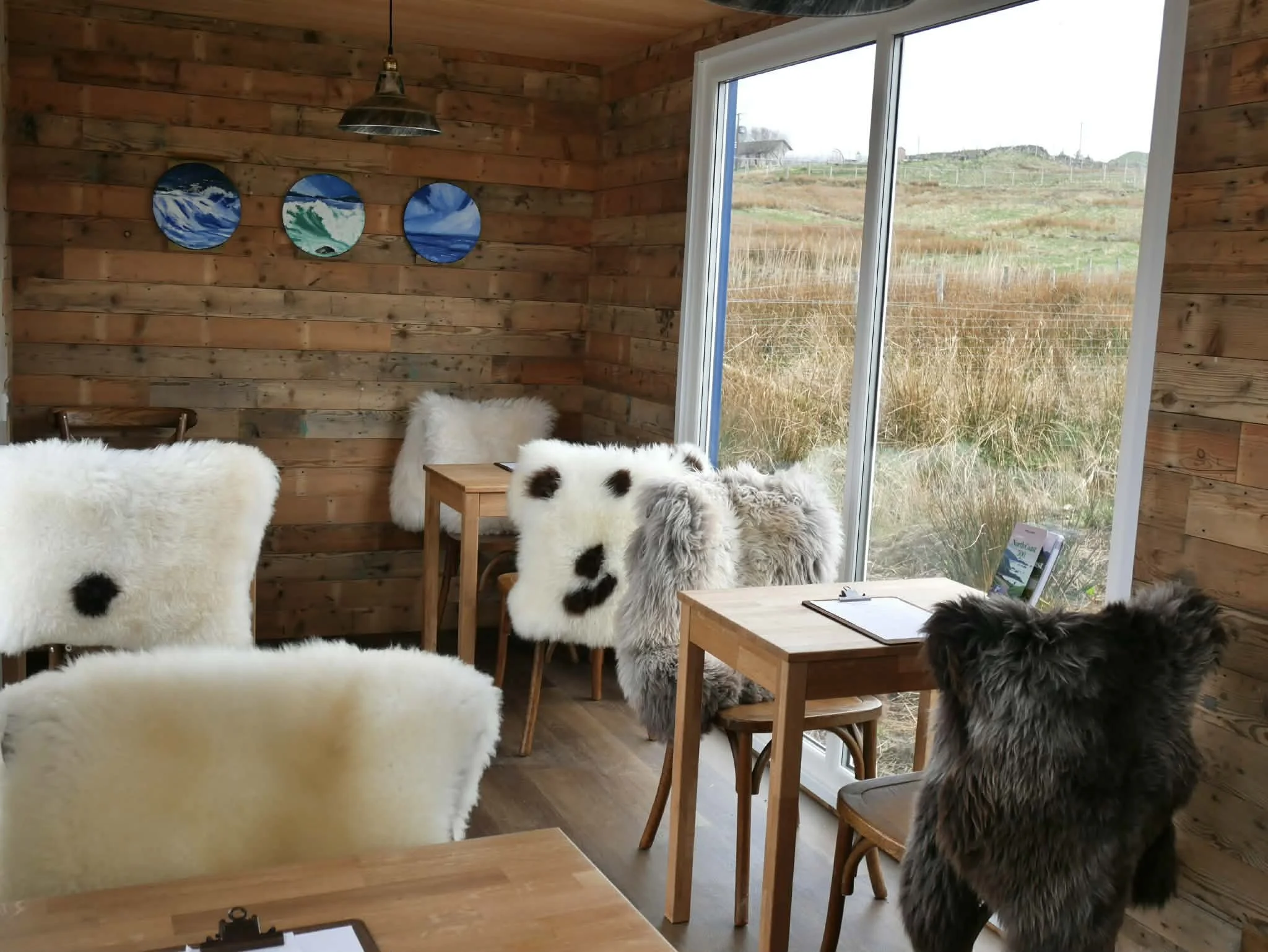 Inside Crofter's Kitchen's new container restaurant — sheepskin-covered chairs, reclaimed timber walls, and floor-to-ceiling windows looking out over the Sutherland moorland. NC500, Scourie.