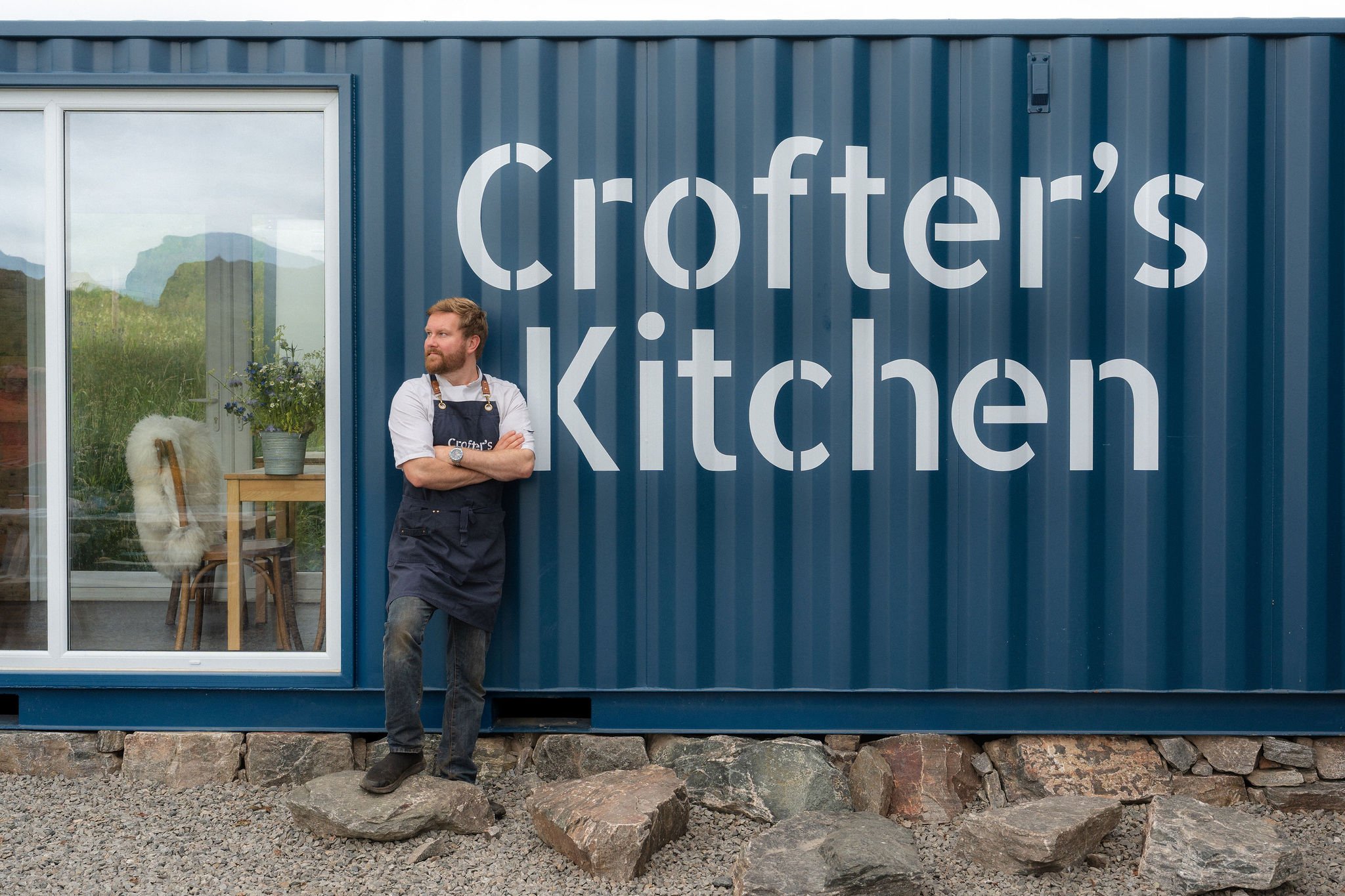 A man with a beard and wearing an apron standing outside a building with a large sign that says 'Crofter's Kitchen'. The building has a large window showing an interior with a table, a potted plant, and a chair. The man is leaning against the building with his arms crossed, looking to the side.