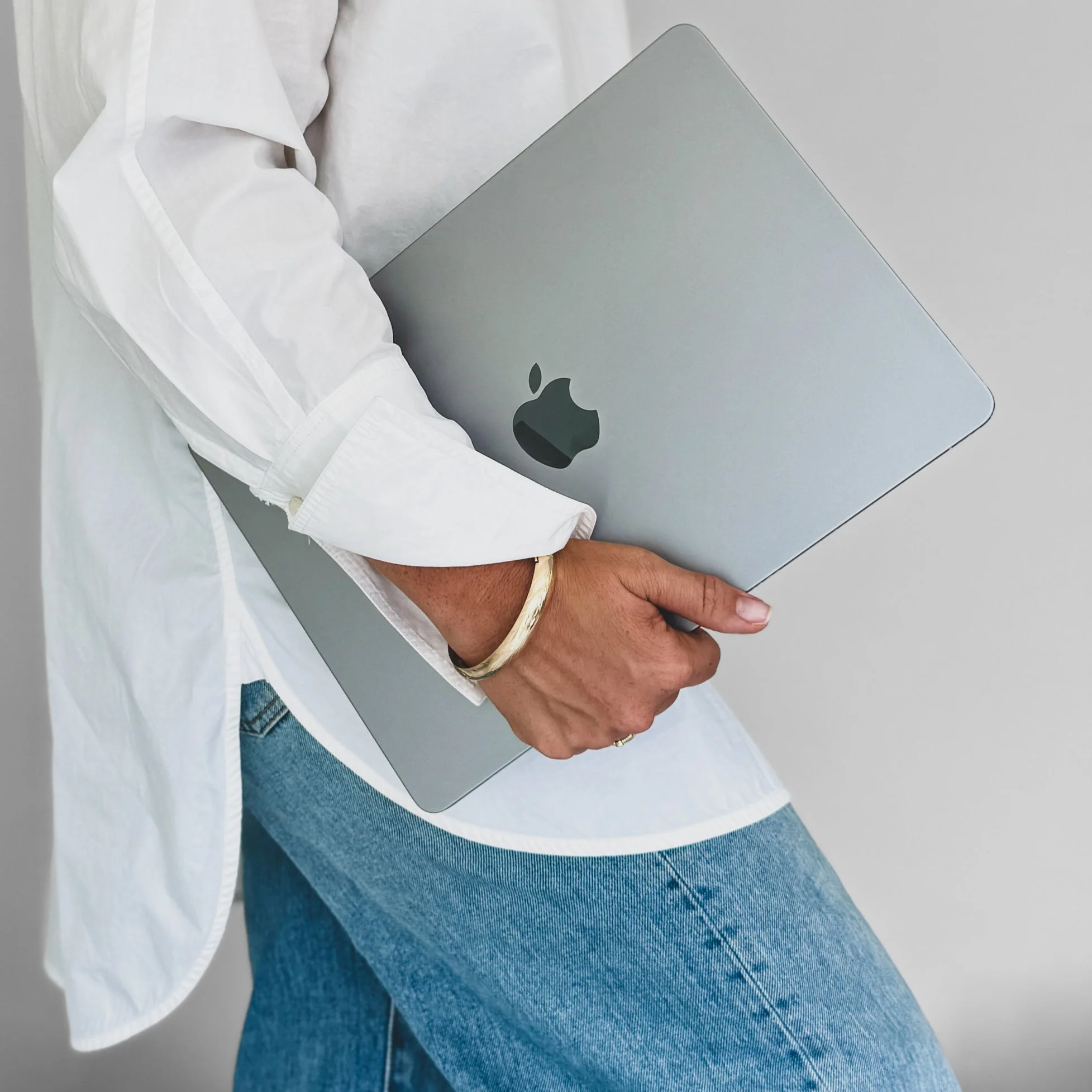 Virtual Personal Stylist, Brianna Albrecht of Begin Anew, wearing white button down shirt over light wash jeans holding laptop in hand wearing gold bracelet