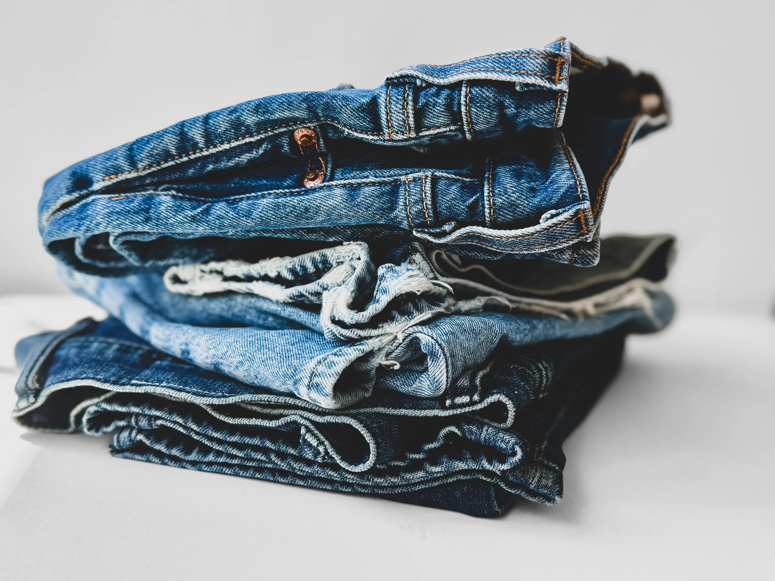 four pairs of folded denim stacked on top of each other with varying shade of blue, distressing set amongst white studio space