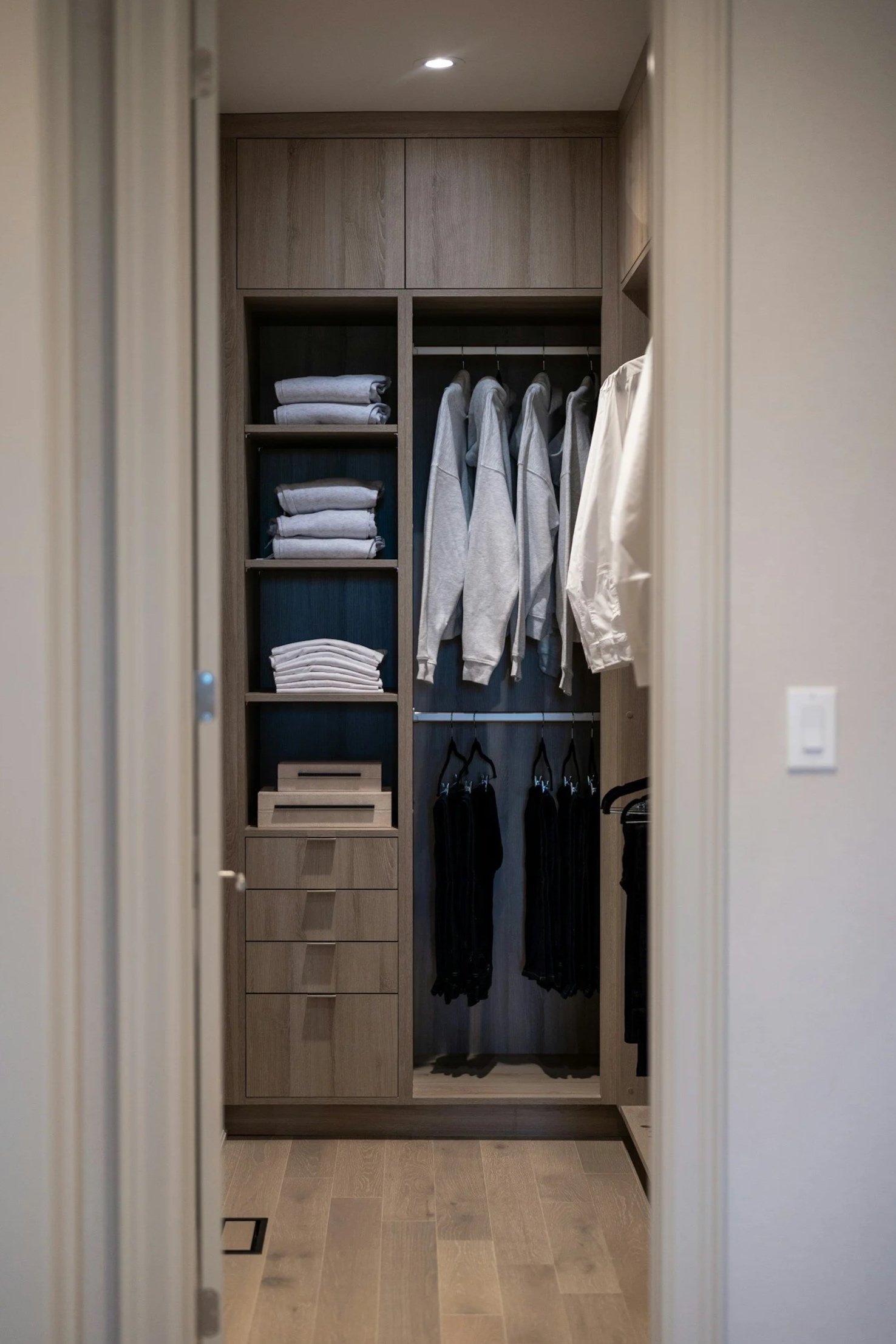 A walk-in closet with built-in shelves and drawers, containing folded clothes, hanging hoodies, and pants, with a wooden floor and neutral-colored walls.