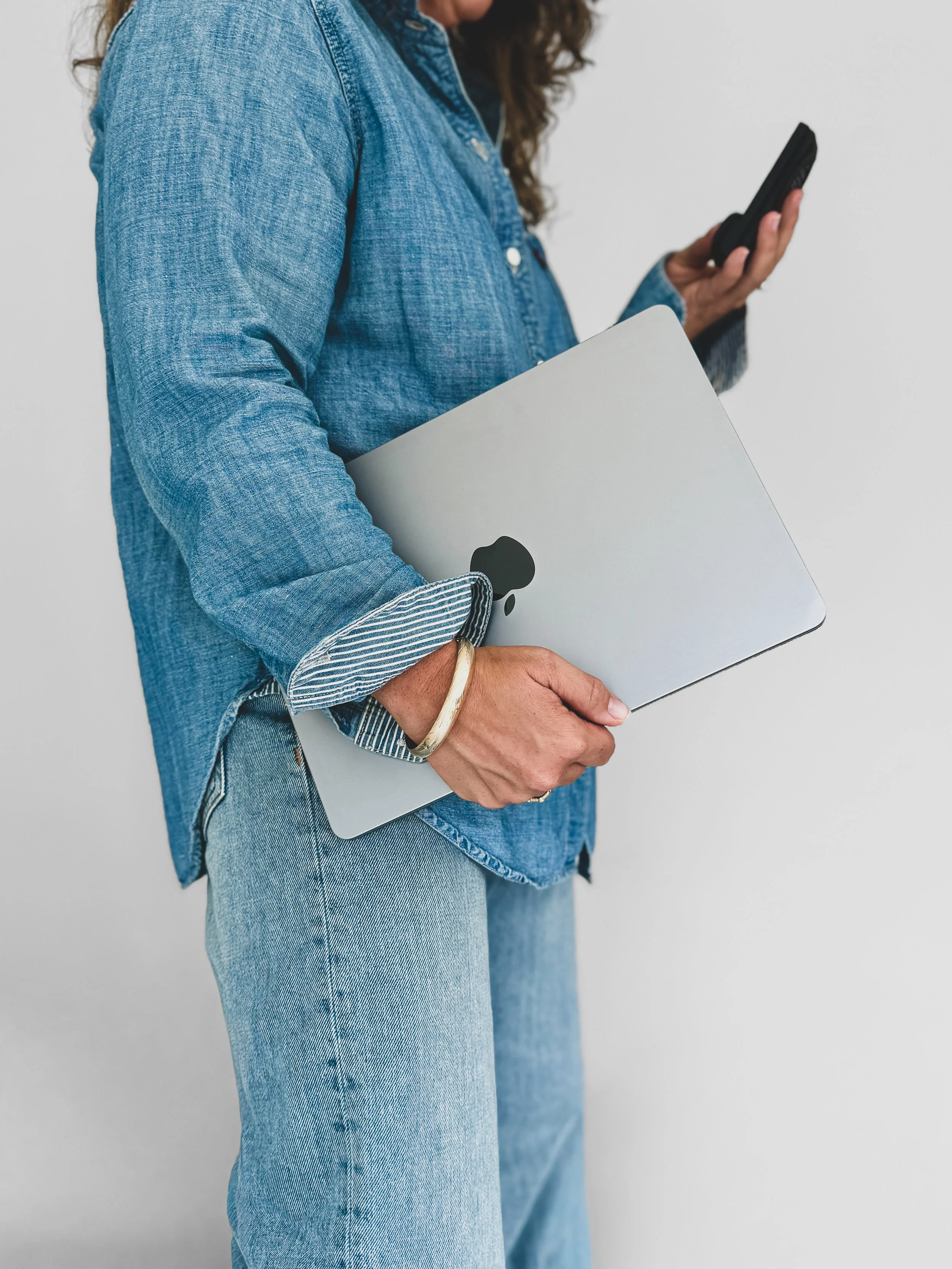 Virtual personal stylist holding laptop in one hand phone in other wearing denim shirt gold bracelet with jeans