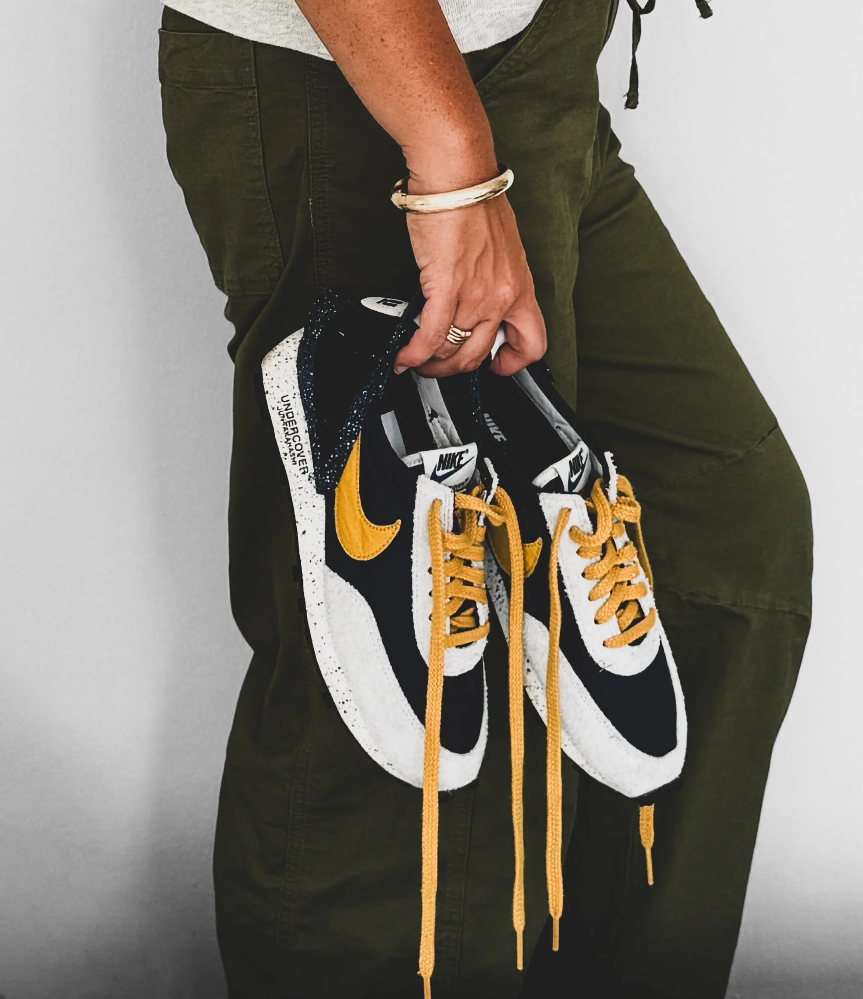 Brianna Albrecht, chicago personal stylist of BEGIN ANEW STYLE holding pair of navy, white, gold Nike Sneakers in right hand along side of leg in dark olive pants. mustard yellow laces hang untied
