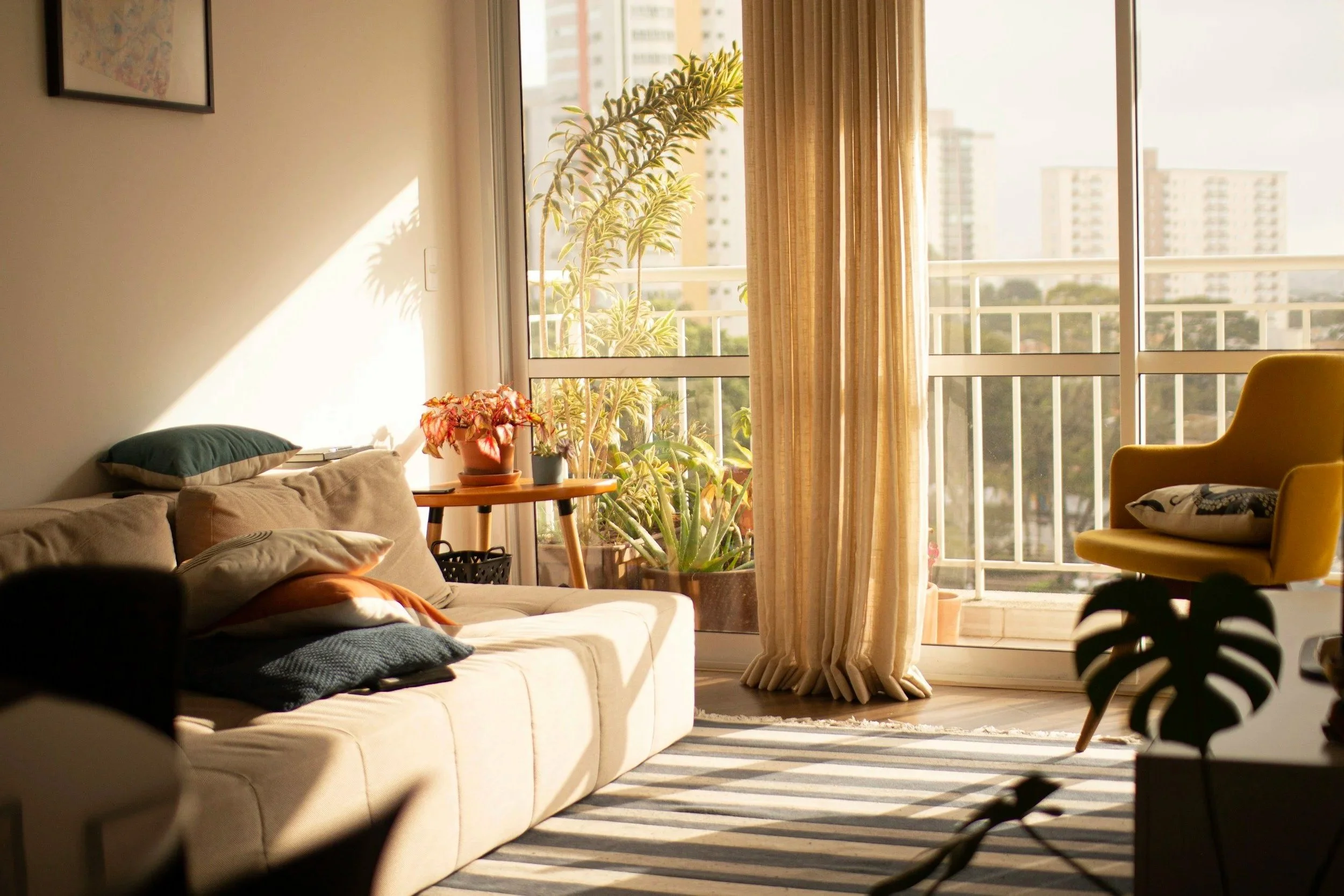 living room with afternoon sun streaming across room onto large cream couch, throw pillows with plant filled balcony in the background