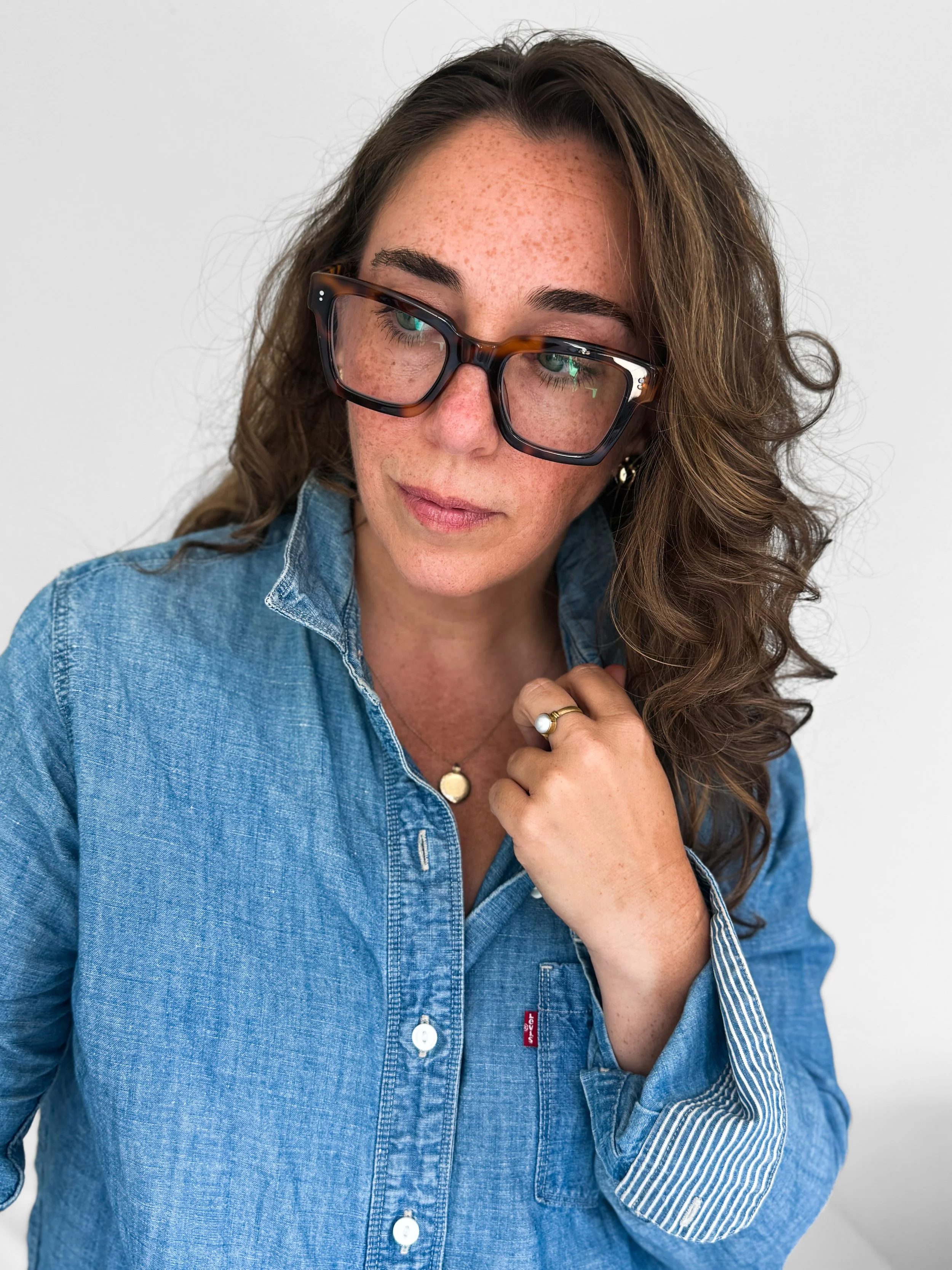 Brianna Albrecht, Chicago personal stylist of BEGIN ANEW STYLE posed with left hand adjusting collar of denim shirt, gold pendant necklace, large frame glasses and light brown wavy hair, looking slightly down out of frame