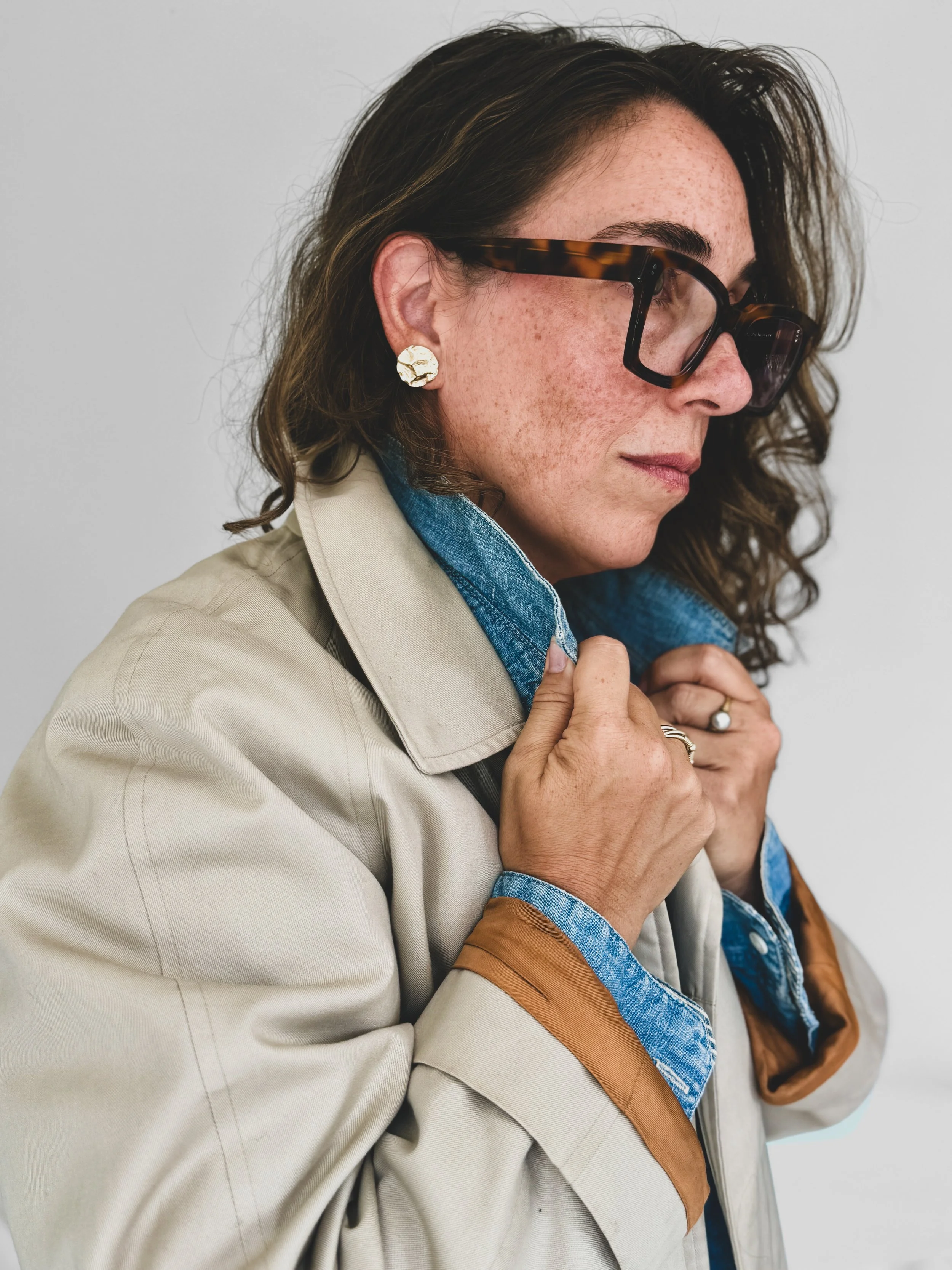 Brianna Albrecht, Chicago stylist of BEGIN ANEW STYLE wearing tan trench coat over denim shirt and dark large frame glasses, holding the shirt collar in both hands standing at profile