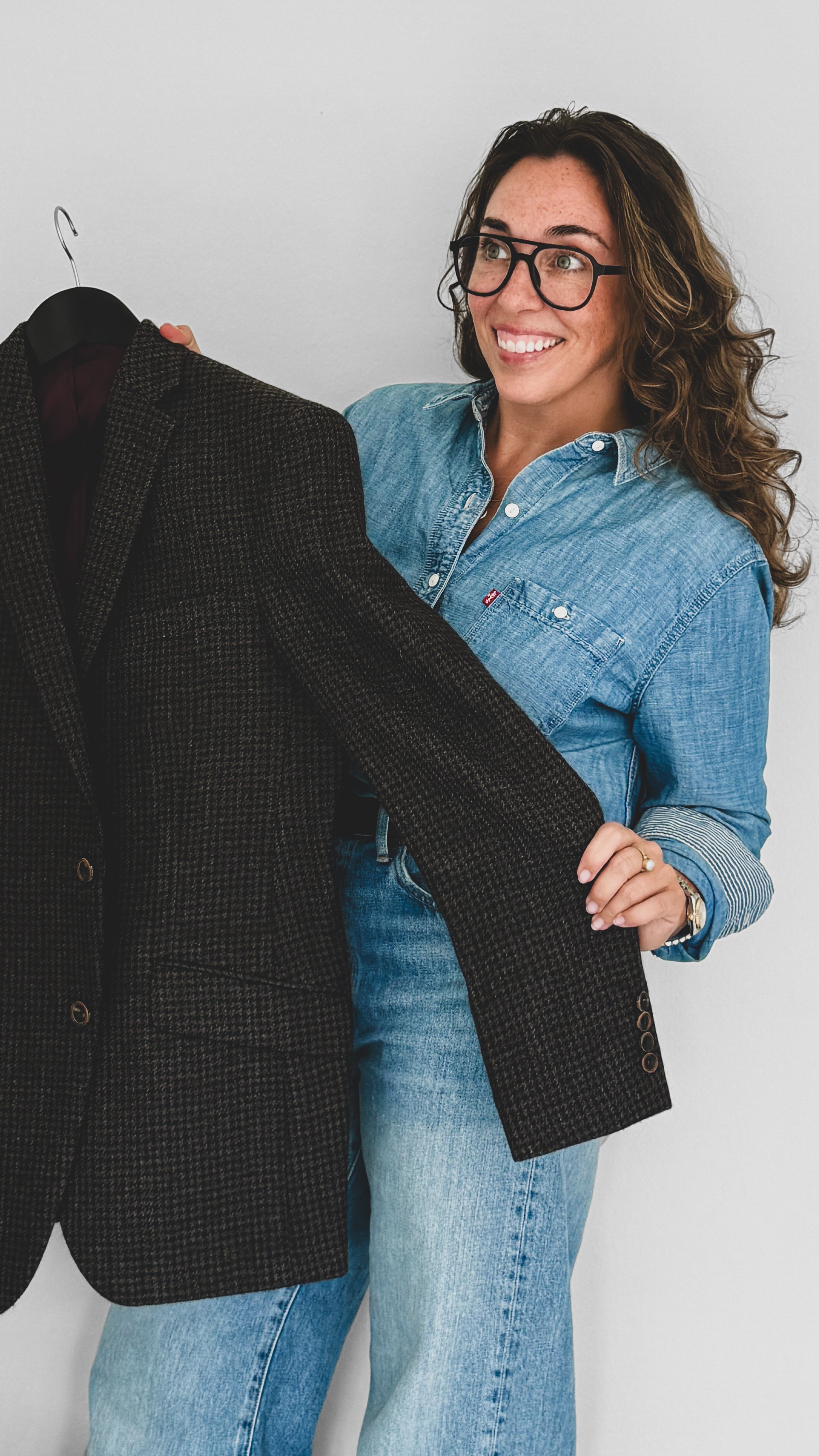 Brianna Albrecht, Chicago Personal Stylist at Begin Anew wearing glasses denim shirt while holding a blazer while smiling at a client out of view