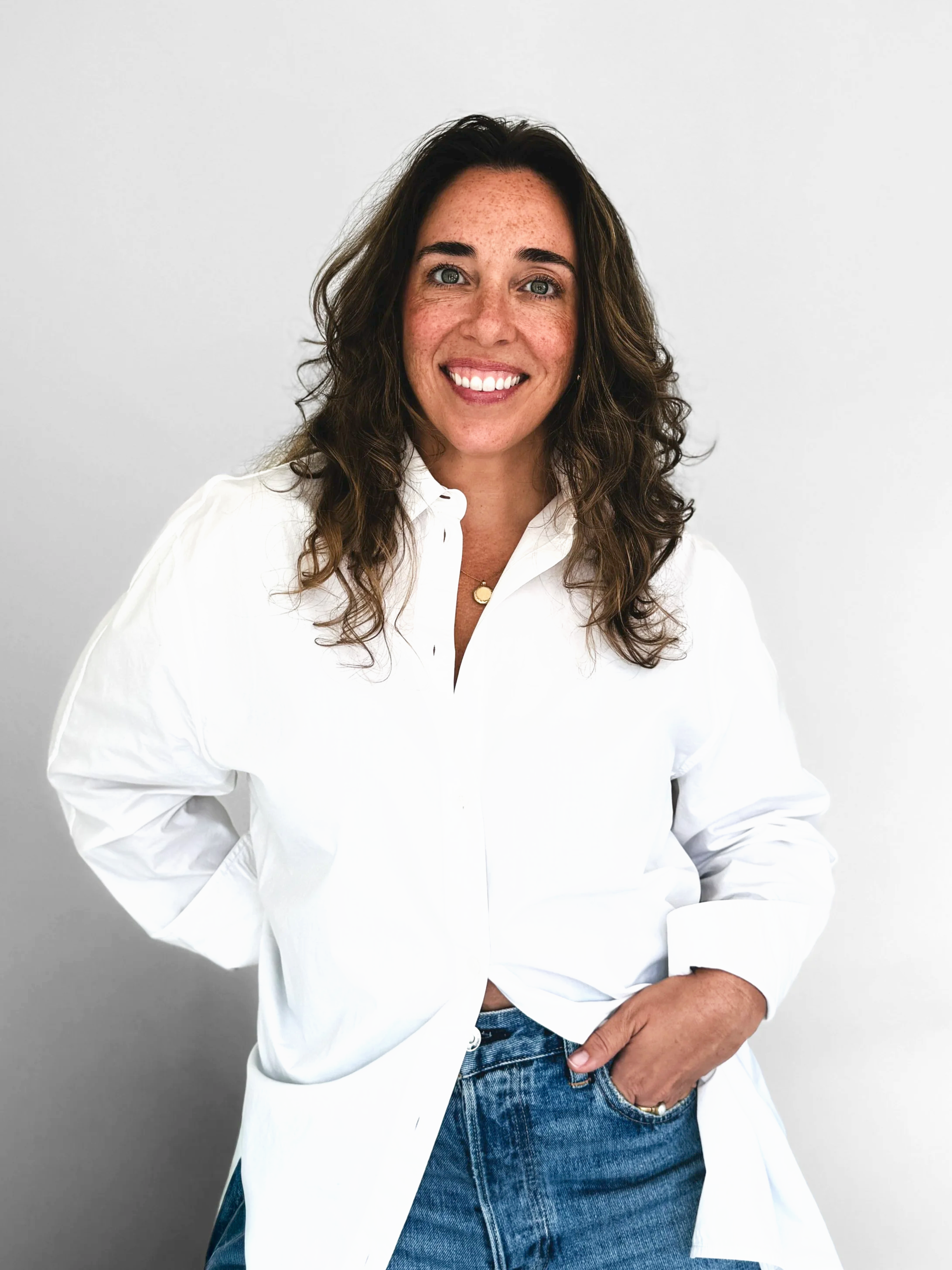 Brianna Albrecht, Chicago Personal Stylist of BEGIN ANEW STYLE smiling directly at camera, wearing white button down with jeans seen with hand in front pocket pulling back part of shirt, wavy hair, gold necklace