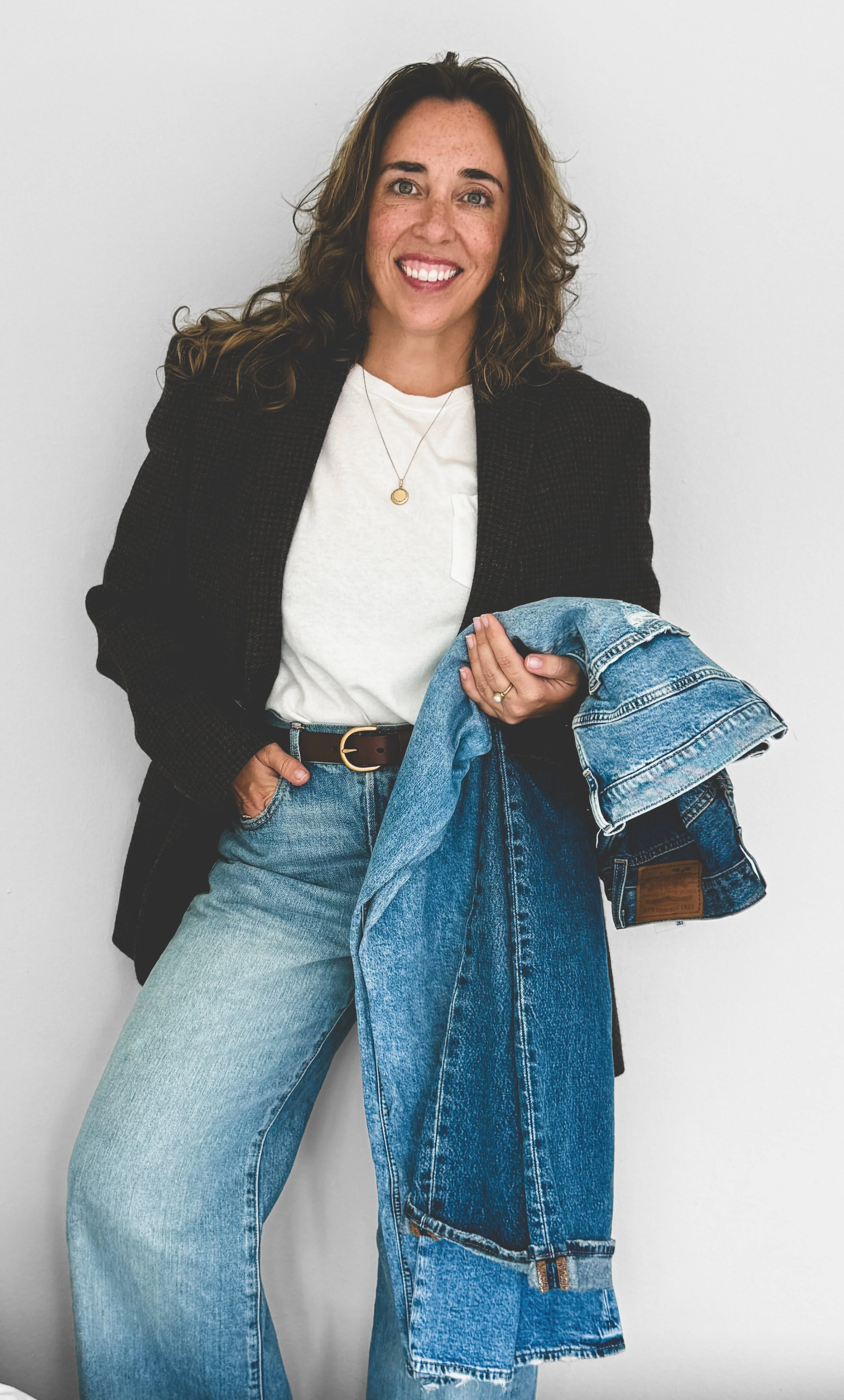 Brianna Albrecht, Chicago Men's Personal Stylist and founder BEGIN ANEW wearing a dark blazer over a white t shirt holding pairs of jeans has curly brown hair