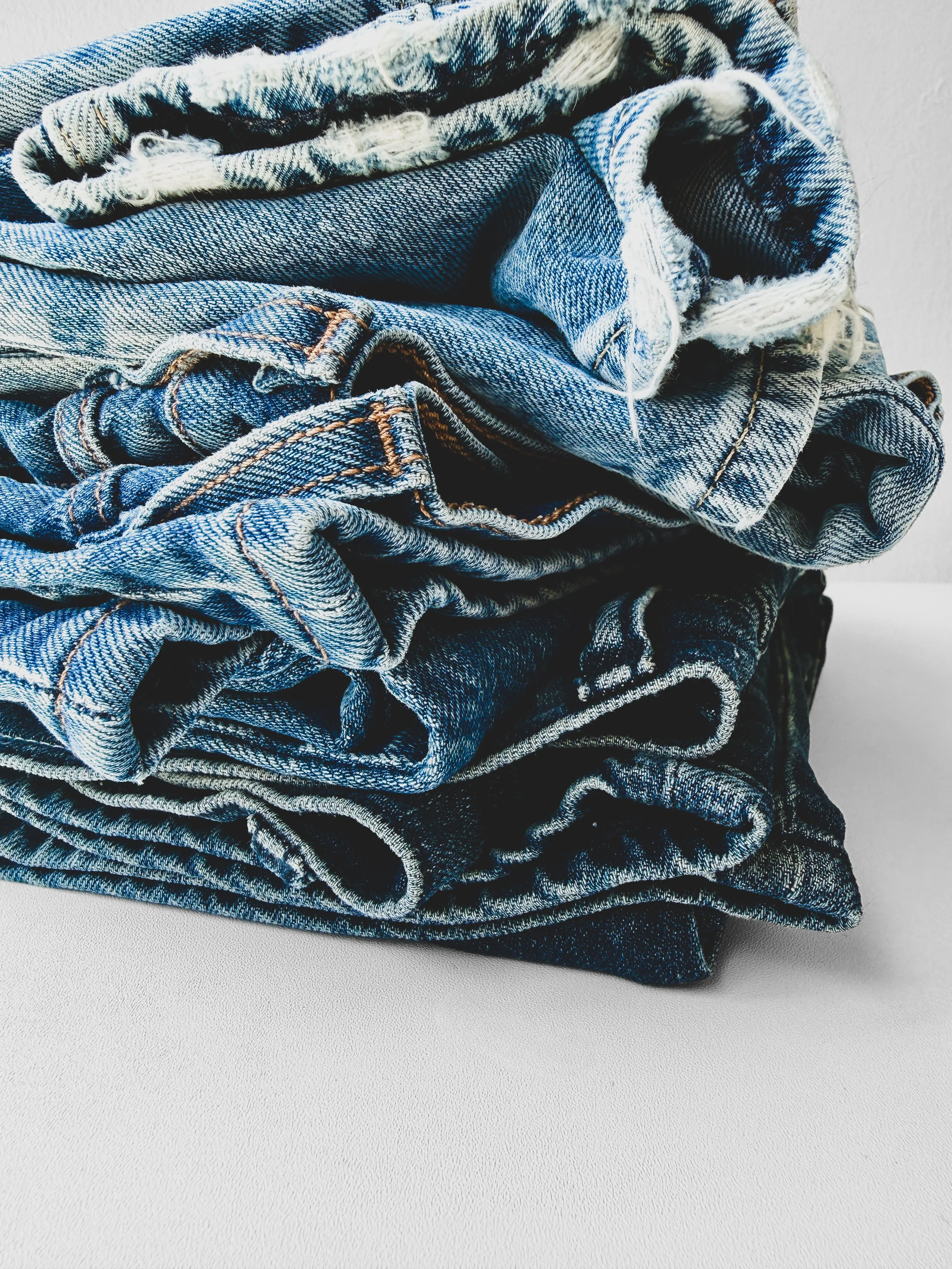 A pile of blue denim jeans with various washes and styles, folded and stacked on a white surface.