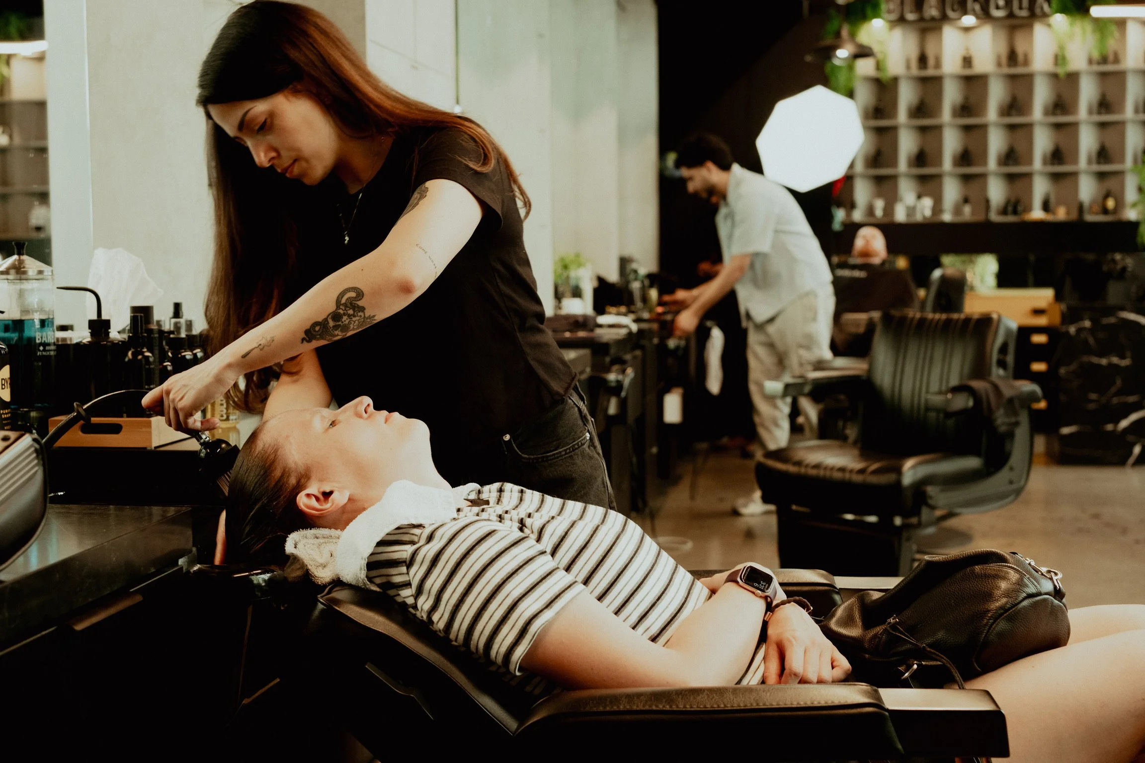 A woman with tattoos on her left arm is lying on a reclining chair in a beauty salon, receiving a facial treatment or skincare procedure. She appears relaxed with her eyes closed, and a woman standing beside her is applying treatment or makeup. The s