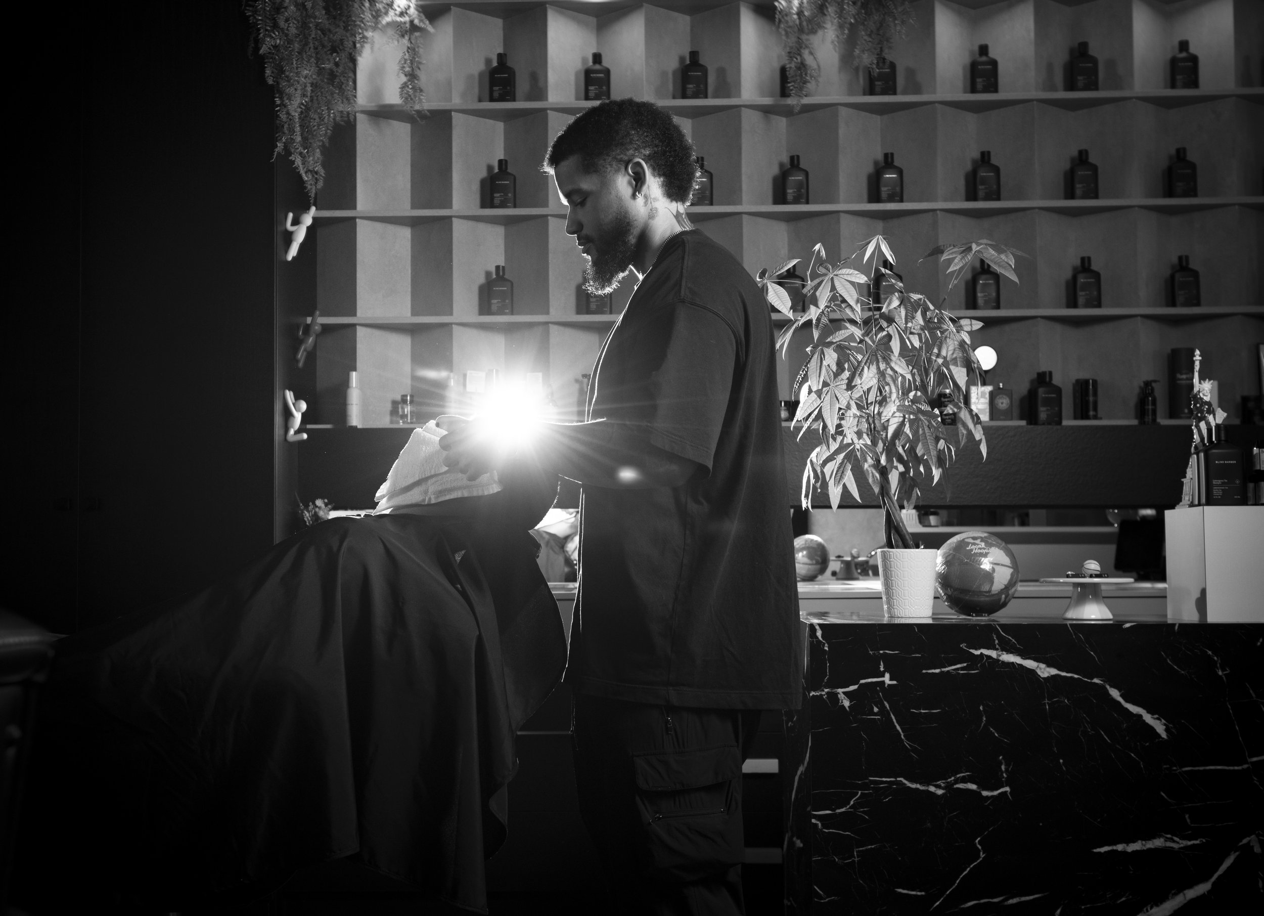 A man getting a haircut in a barbershop, with shelves of bottles behind him and plants in the background.