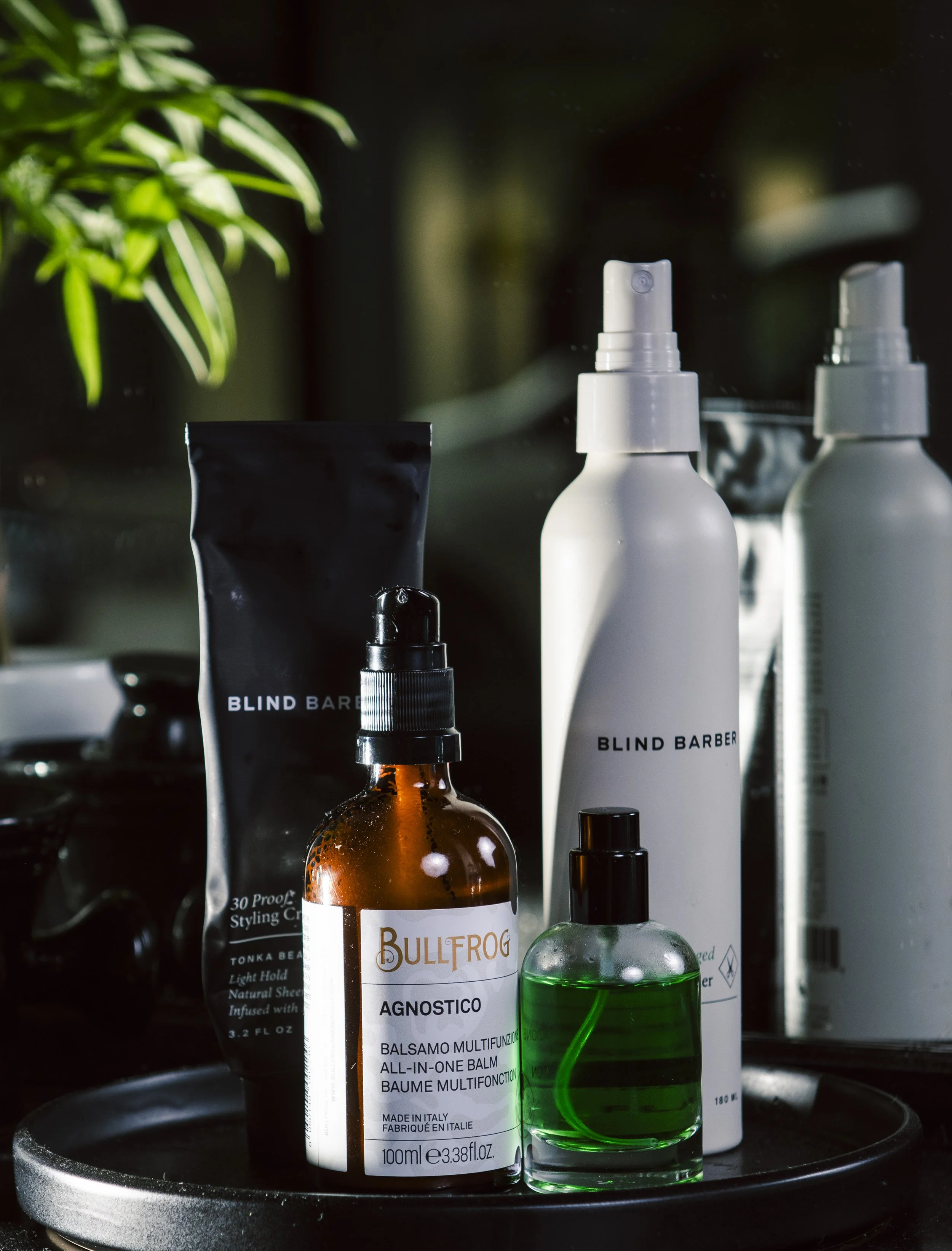 Assorted hair styling products including a white bottle labeled 'BullFrog Agnostico', a green spray bottle, and other containers, with a green plant in the background, placed on a black tray.