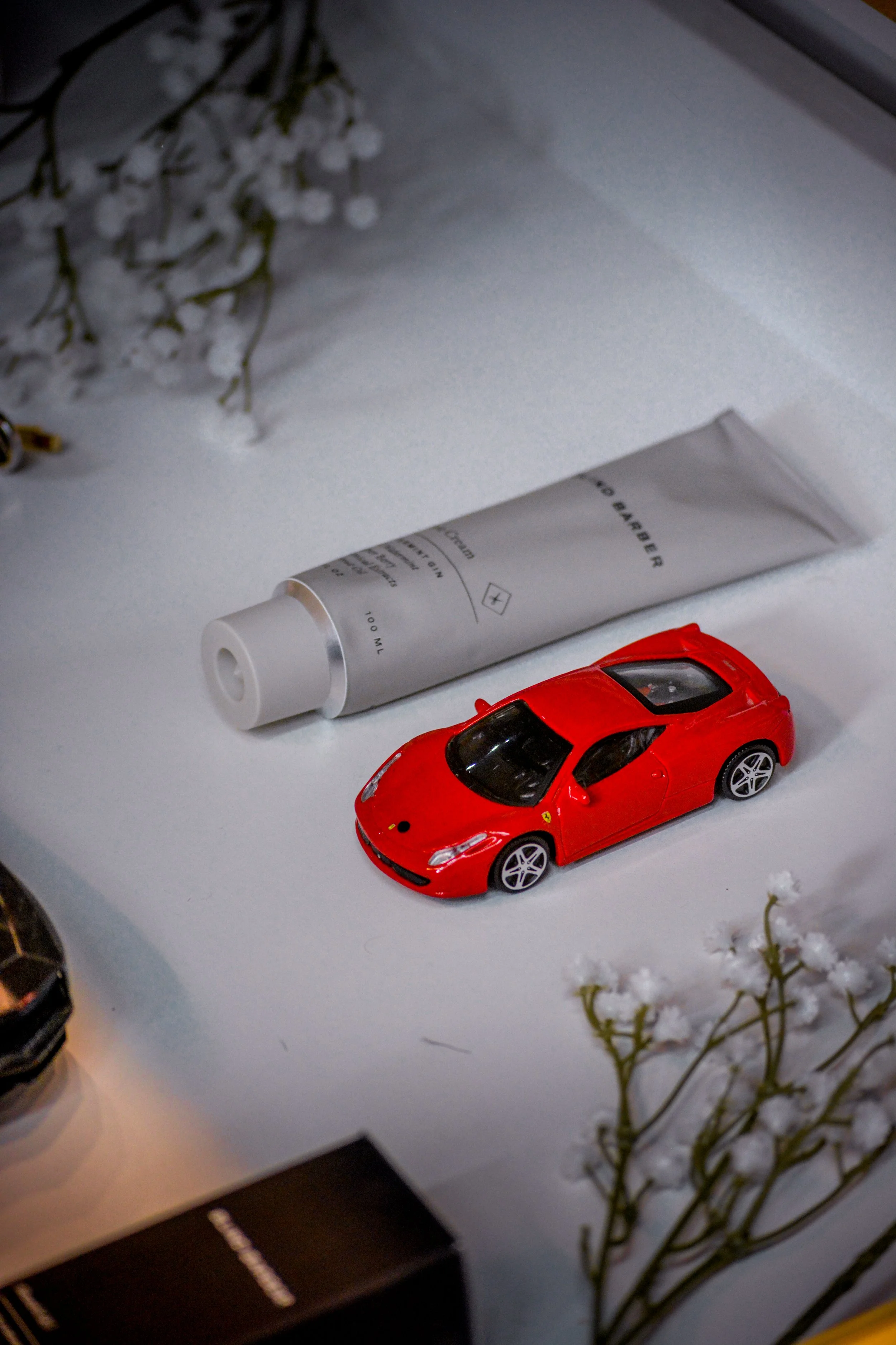 A red toy sports car, a tube of skincare product, and white flowers on a white surface.