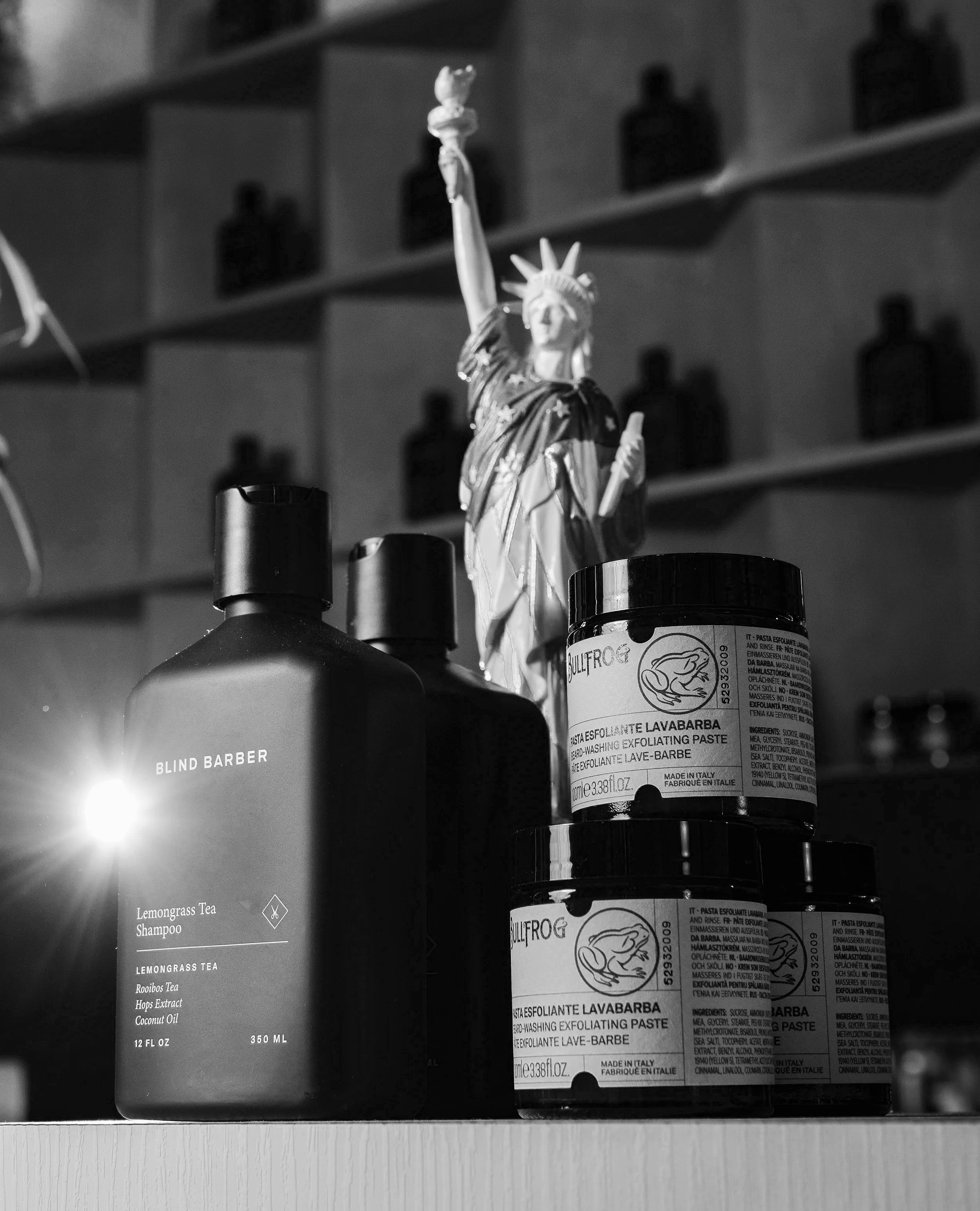 Black and white photo of a shelf with bottles and a small statue of the Statue of Liberty on top of a bottle. The label on the bottle reads "LAVABARBA" and "exfoliating paste."