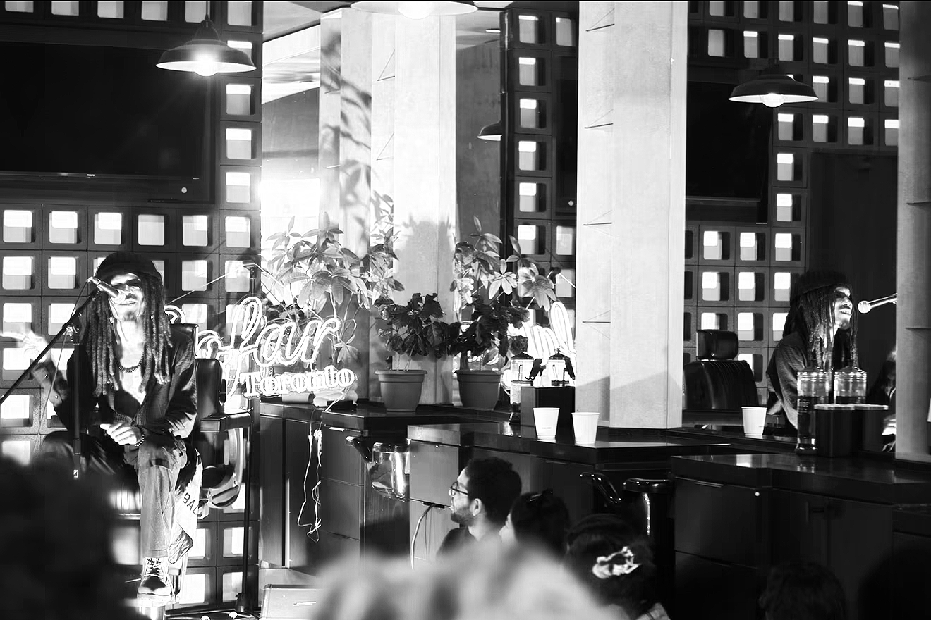 A black and white photo of a music performance in a modern indoor venue. Two musicians with dreadlocks, wearing dark clothing and hats, are seen singing into microphones. The stage features potted plants, a neon sign that reads 'Bar Toronto,' and con