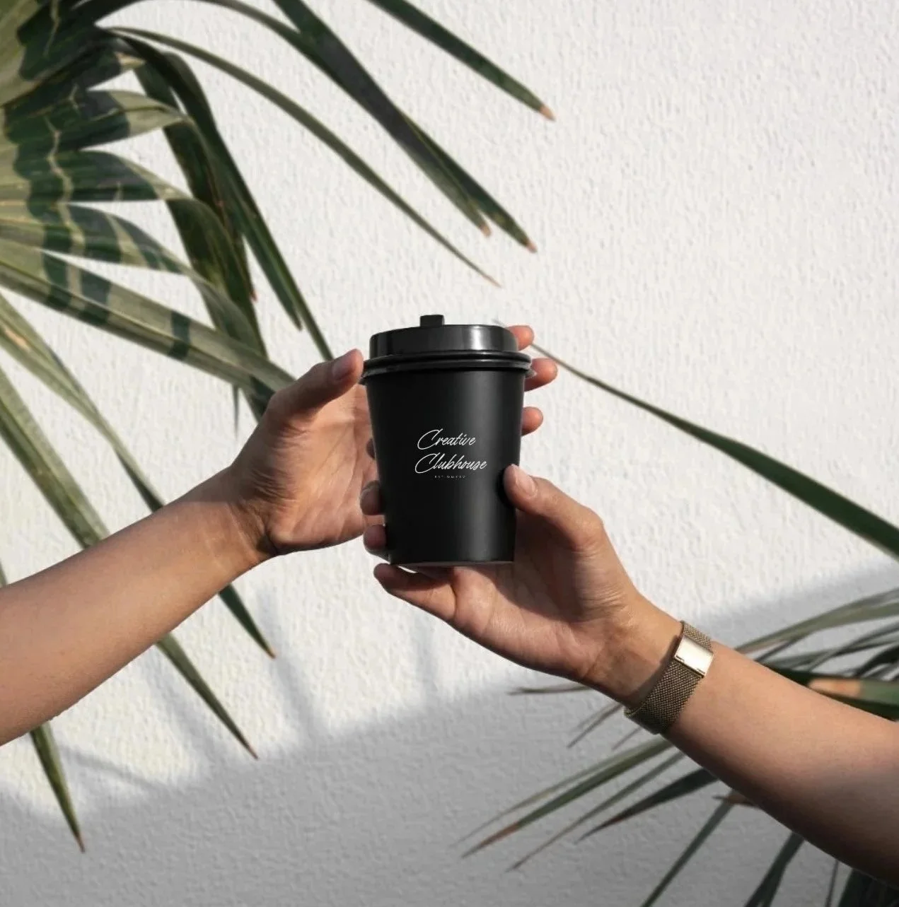 Two hands holding a black coffee cup with a lid, with the words 'Creative Clubhouse' written on it, against a backdrop of large green leaves and a white textured wall.