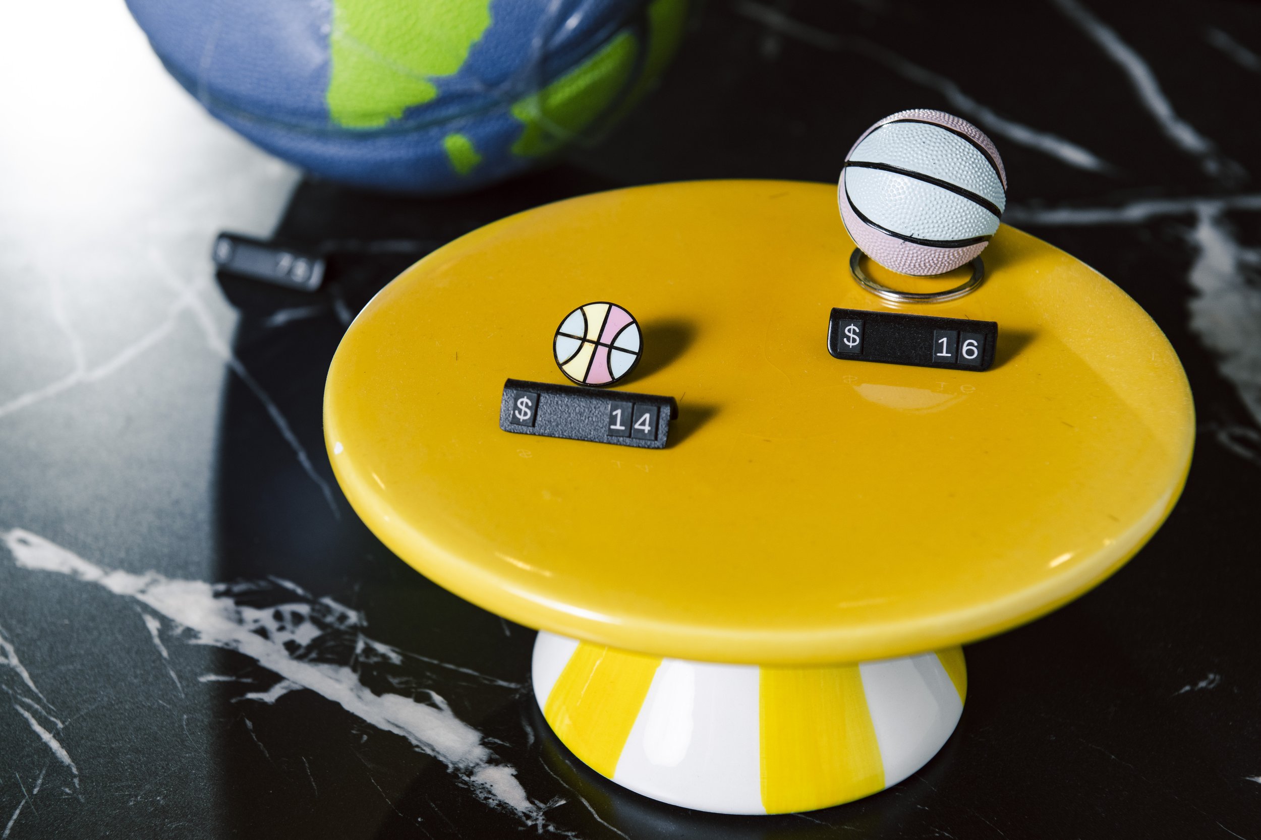 Close-up of a yellow side table with a basketball-shaped keychain, a basketball logo, and price tags, with a partly visible globe in the background and black marble surface underneath.