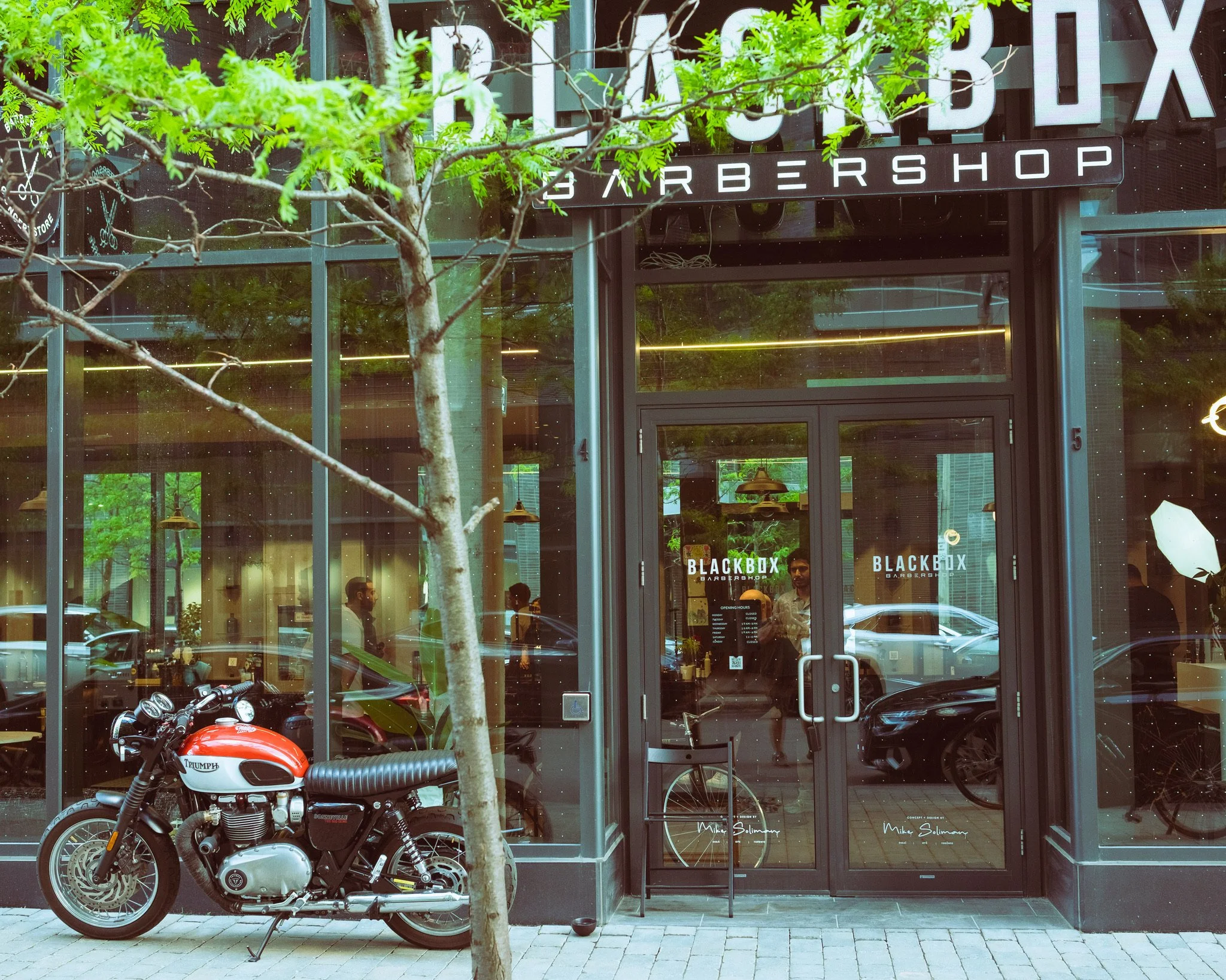 A storefront with large glass windows and a sign that reads 'BLACKBOX BARBERSHOP'. In front of the shop, there is a parked red and black Triumph motorcycle and a small tree. The interior of the shop is visible through the windows, showing people insi