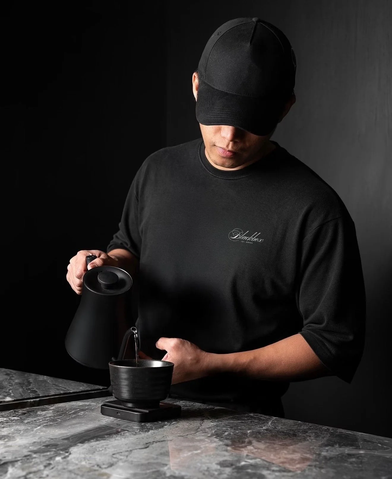 A man dressed in black wearing a cap, pouring hot water from a kettle into a black bowl on a kitchen scale, with a dark background.