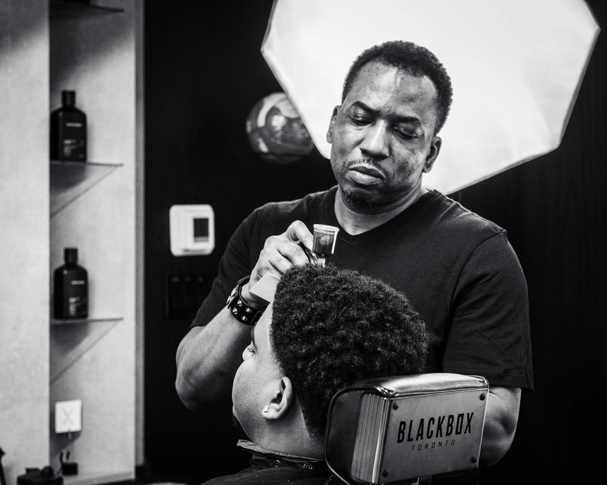 A barber giving a haircut to a young boy in a barbershop. The barber is holding a pair of scissors and a spray bottle, and is focused on trimming the boy’s hair. The boy’s head is slightly bowed, and he appears relaxed. There is a shelf with hair pro