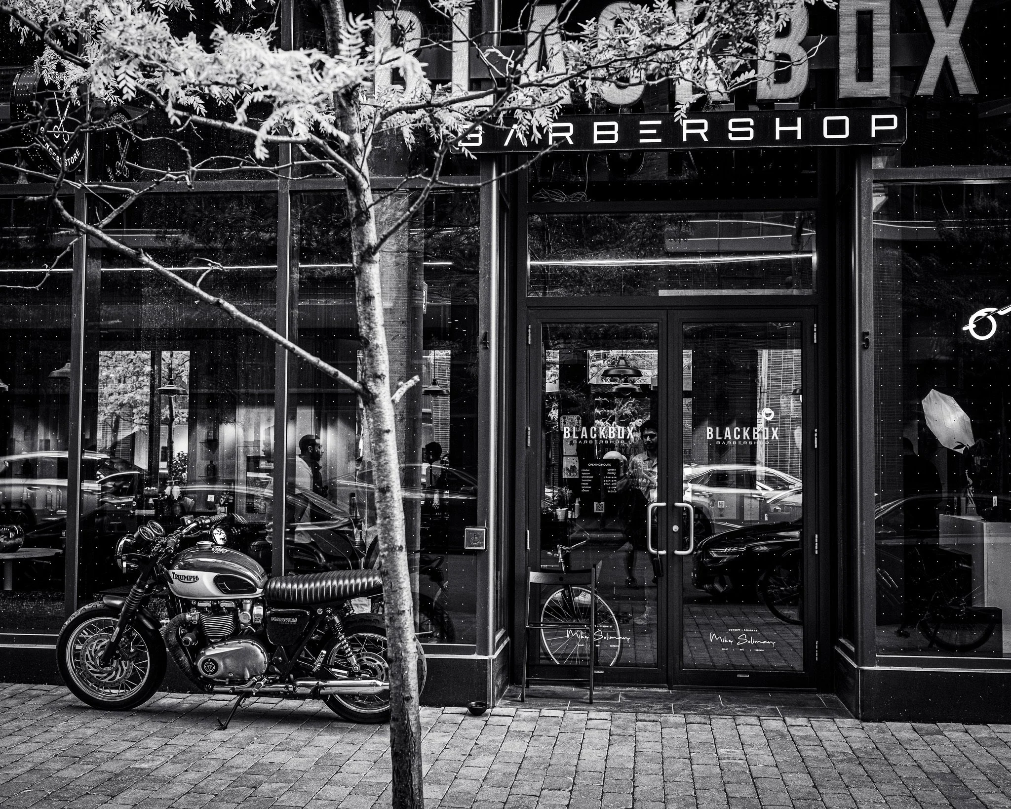 Black and white photo of a motorcycle parked outside a barber shop. The shop has large glass windows with people visible inside. There is a tree in the foreground and a sign for the barber shop above the entrance.