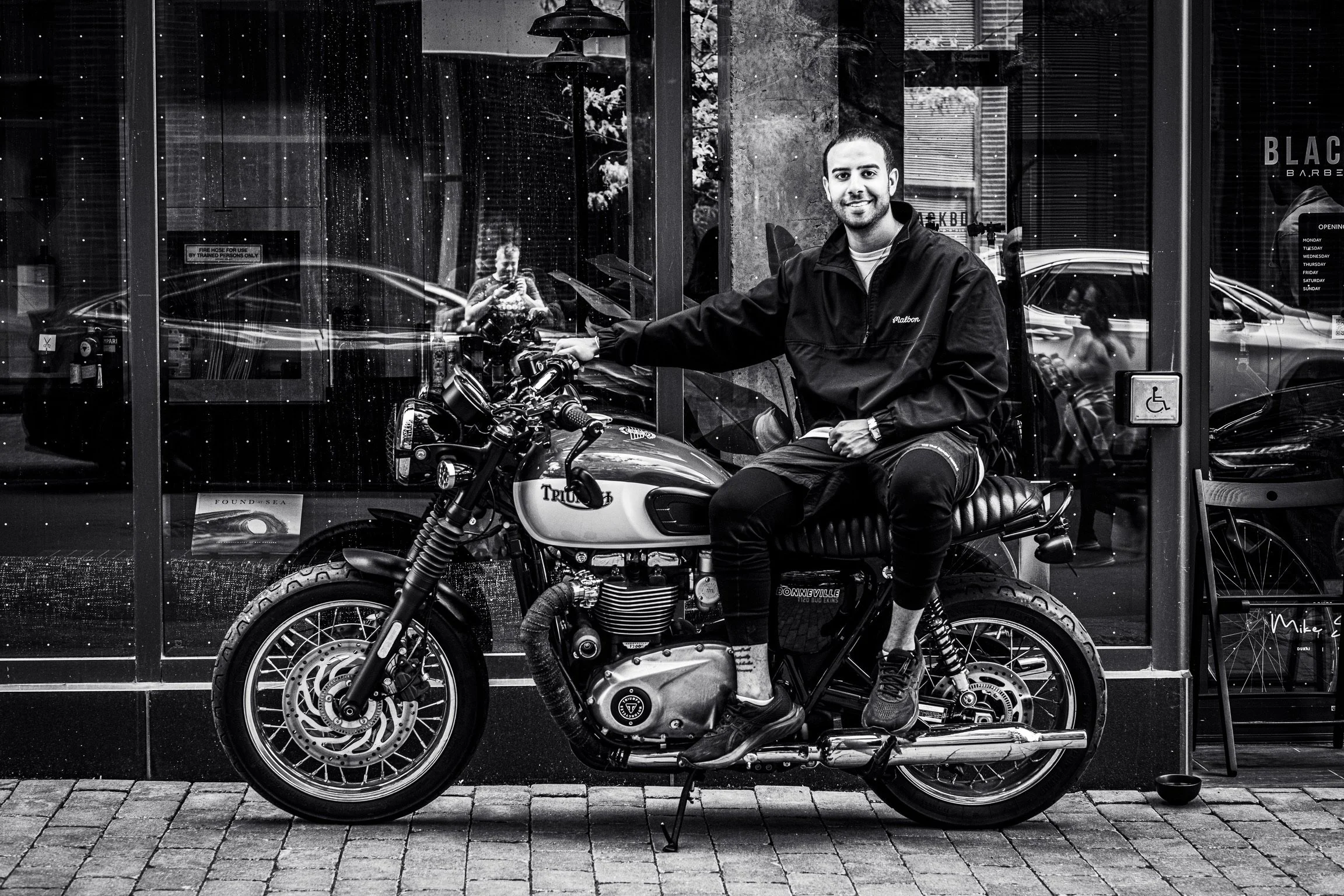 A smiling man sitting on a Triumph motorcycle parked outside a storefront with large glass windows. The reflection of a woman taking the photo and other pedestrians can be seen in the glass.